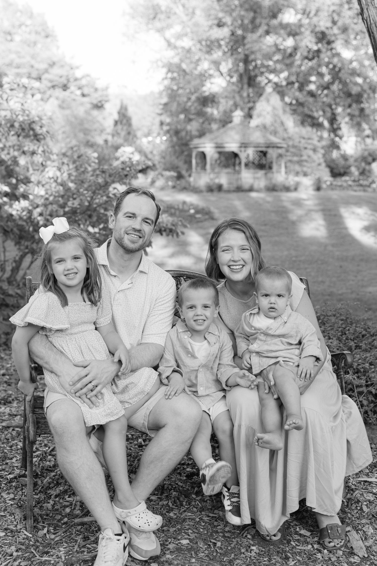 Black and white outdoor portrait of a family of five seated together in a garden with a gazebo in the background.
