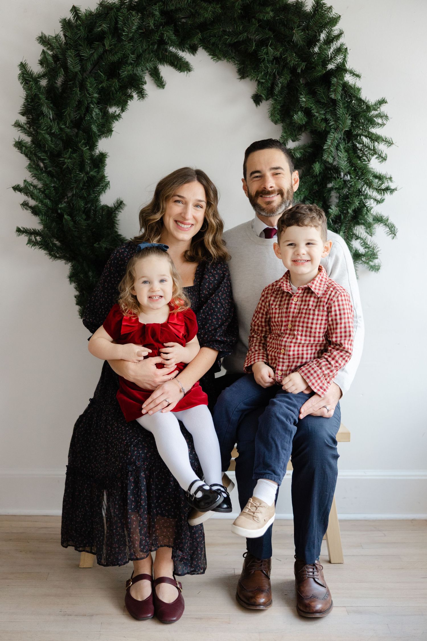 A family sitting together against a white wall with evergreen garland, wearing coordinated holiday outfits.