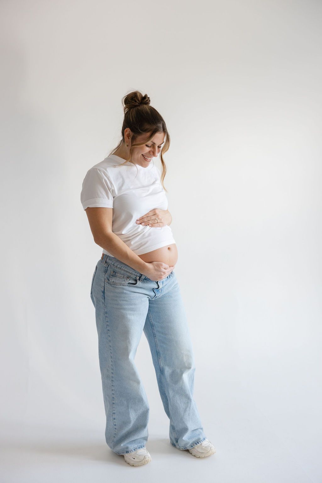 Pregnant person in white t-shirt and light blue jeans poses against white background showing growing baby bump.
