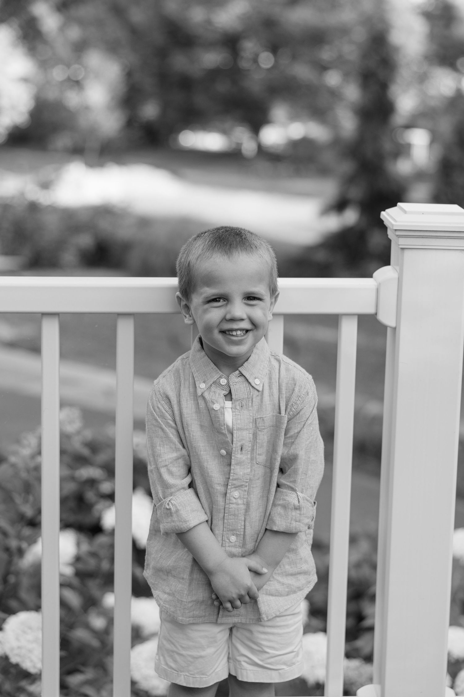 A young child in a light colored shirt smiles while standing against a white porch railing outdoors.
