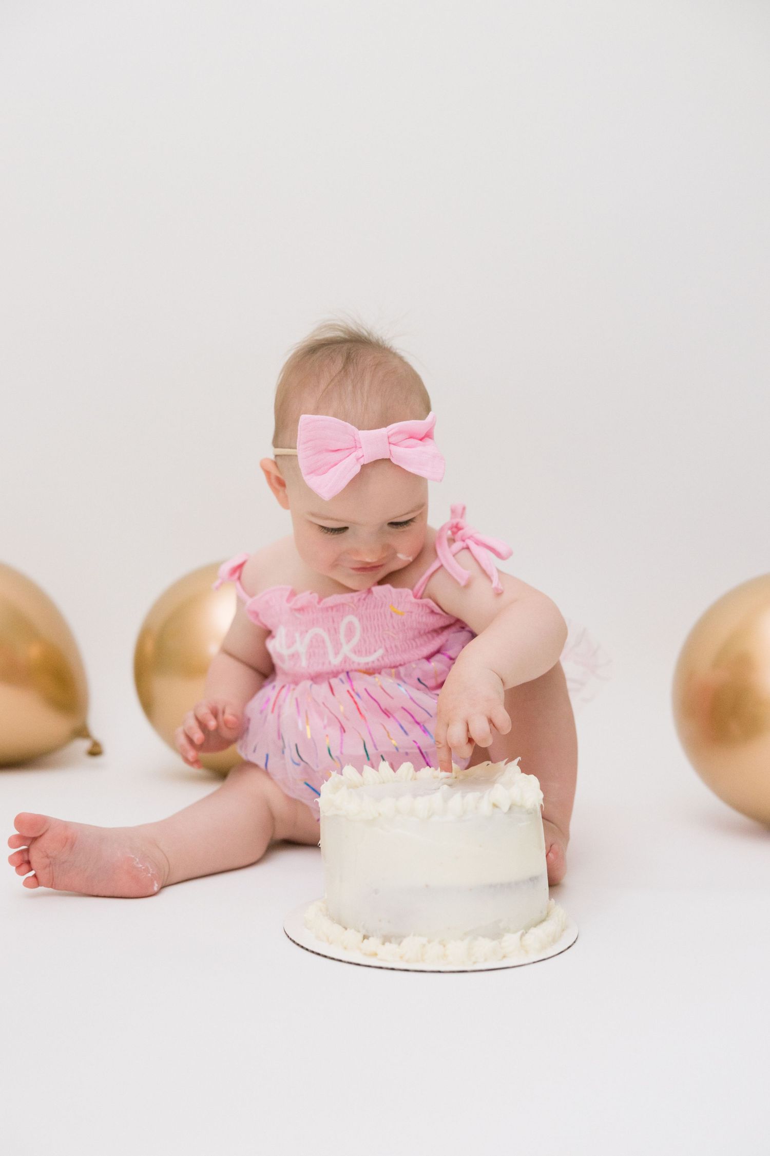 Baby wearing pink outfit reaching for white birthday cake during first birthday cake smash photoshoot.