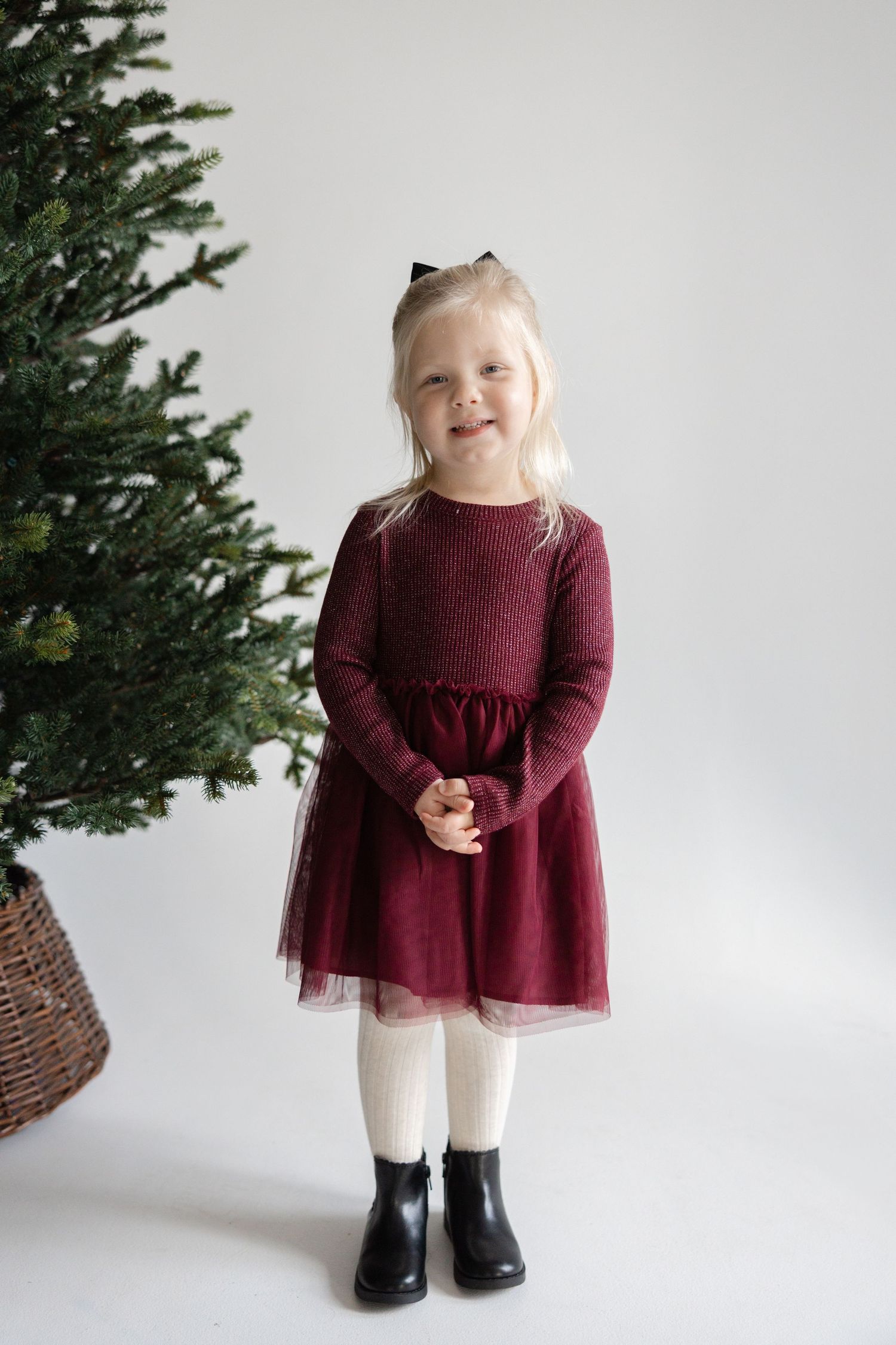 A small child in a burgundy dress and black boots poses against a white backdrop near a Christmas tree.