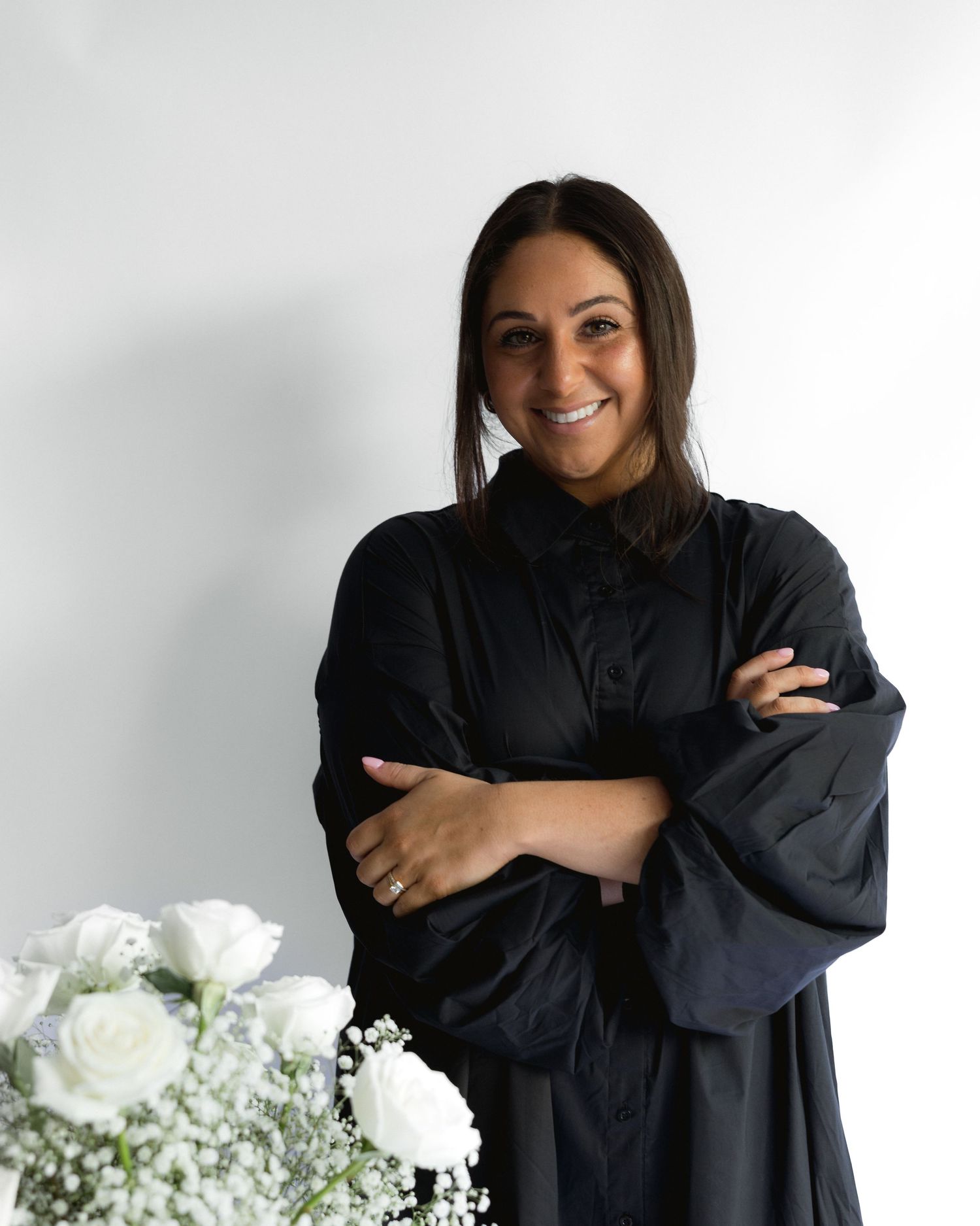 Professional portrait of someone wearing a black graduation gown standing next to white flowers against a light background.
