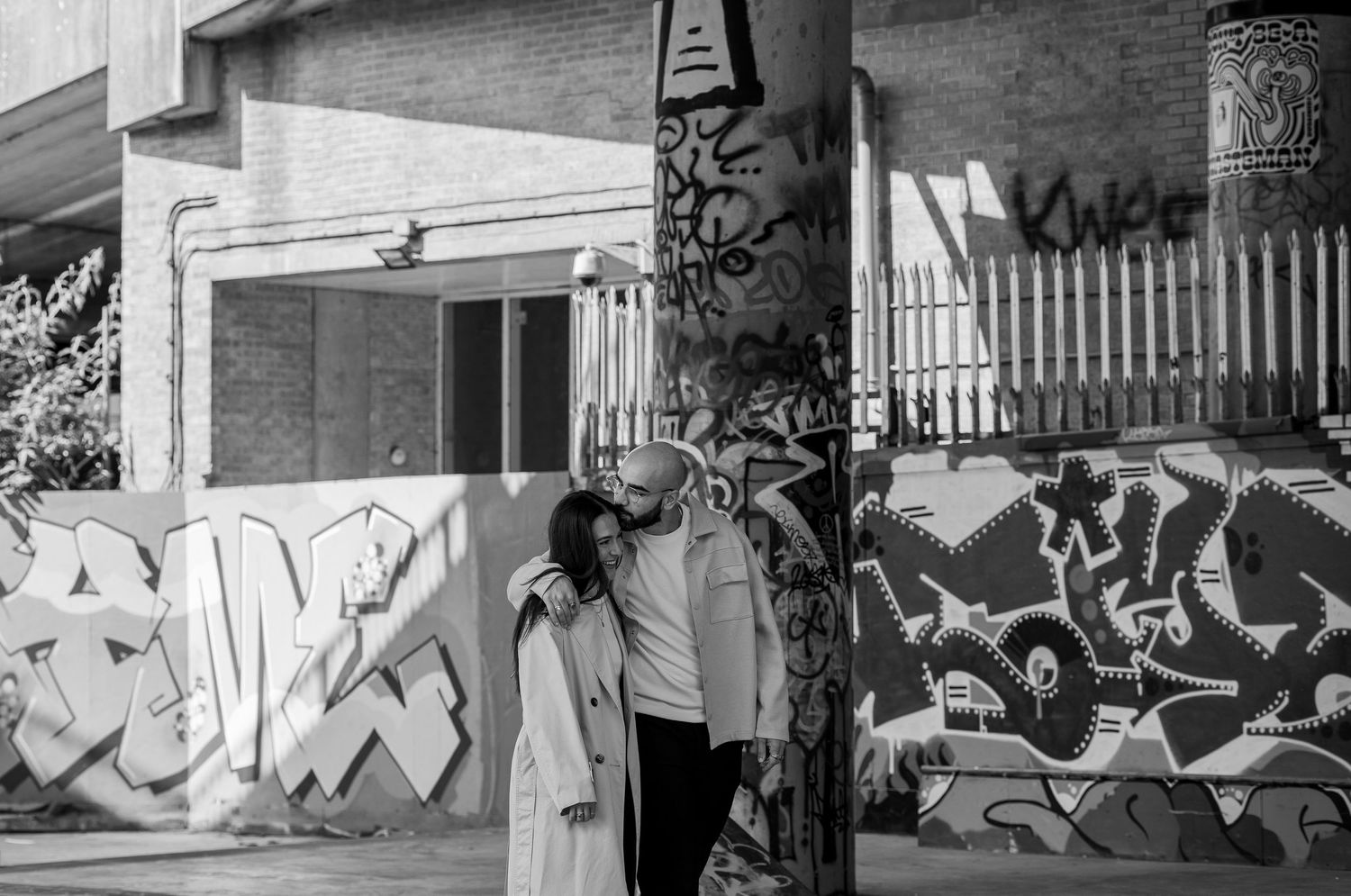 Couple walking with graffiti behind them. He is kissing her on the cheek and she is giggling. Natural documentary wedding photography