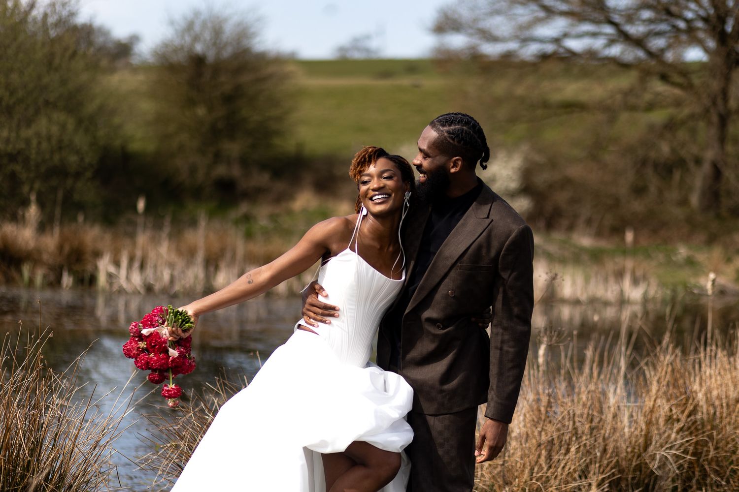 Couple captured candidly hand in hand by a serene lake, carrying a bridal bouquet. Natural documentary wedding photography