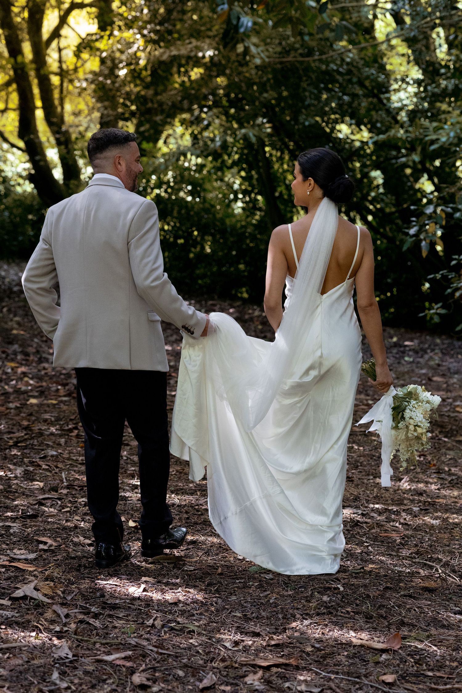 Couple captured walking away with groom holding the brides dress up in a hazy forest. Natural documentary wedding photography