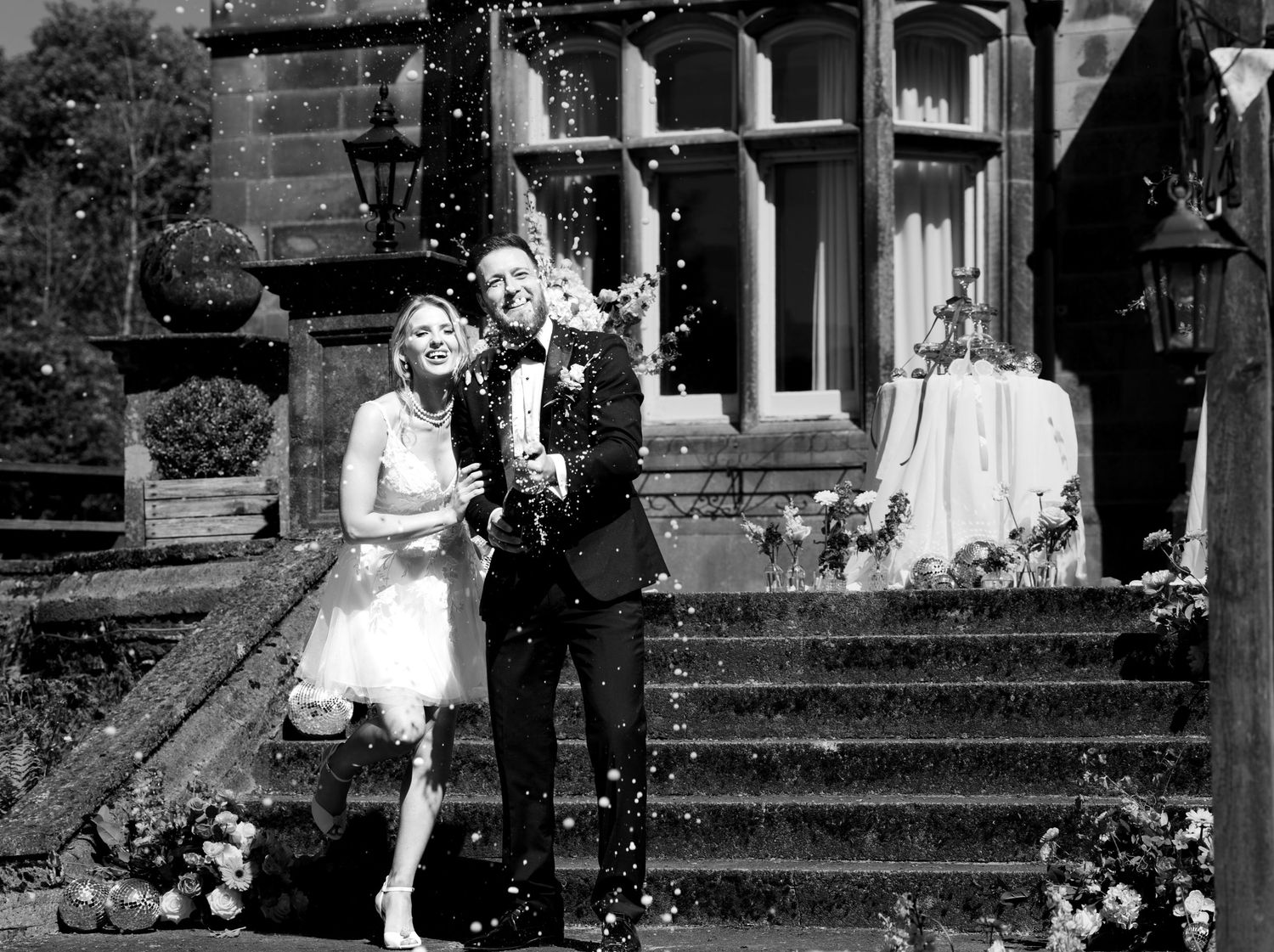Joyous couple on the steps outside their venue.  popping champagne. Big smiles. Natural documentary wedding photography. Black and white