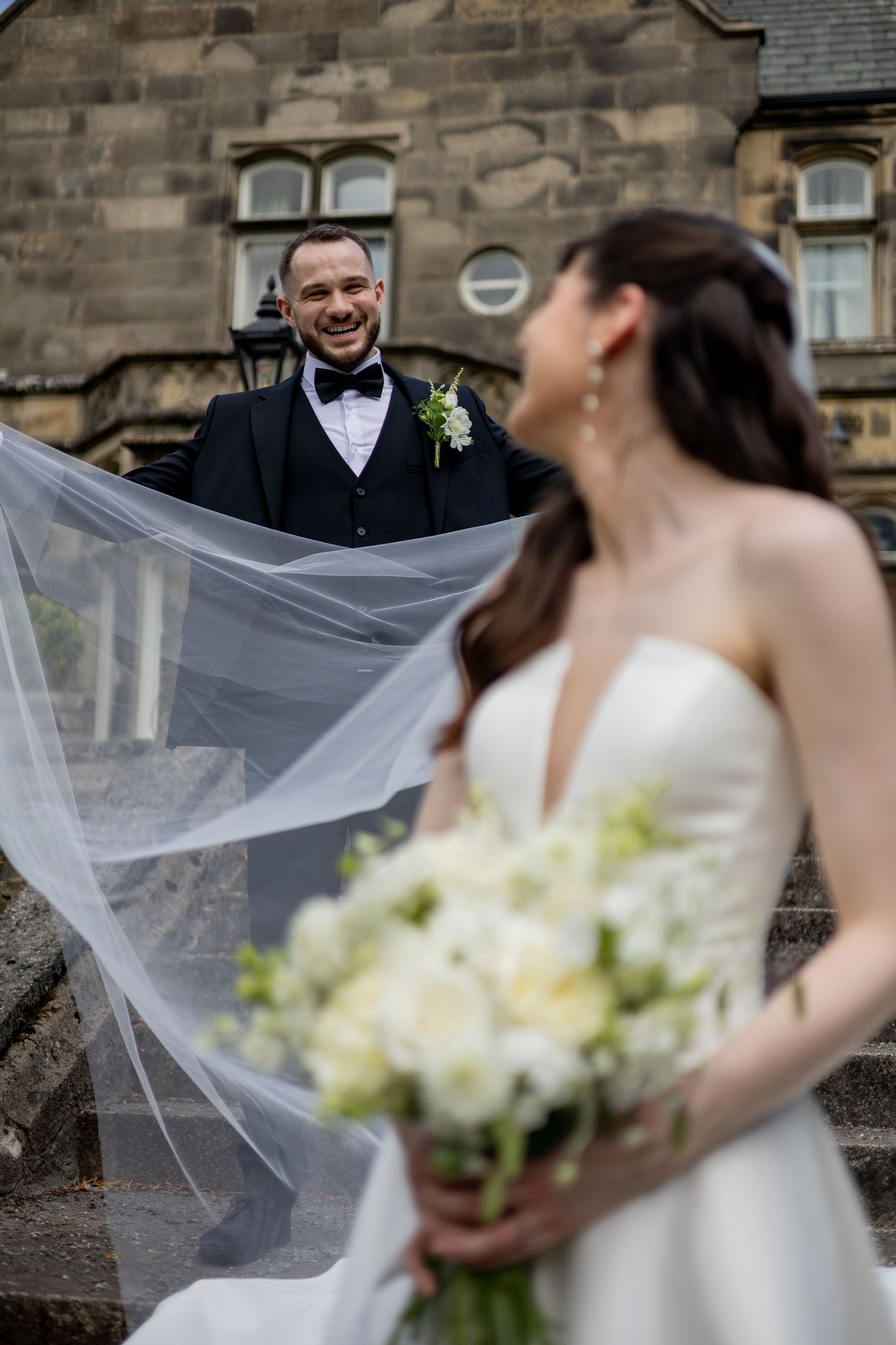 Joyous couple on the steps outside Hargate Hall. Groom holding the brides long veil. Big smiles. Natural documentary wedding photography