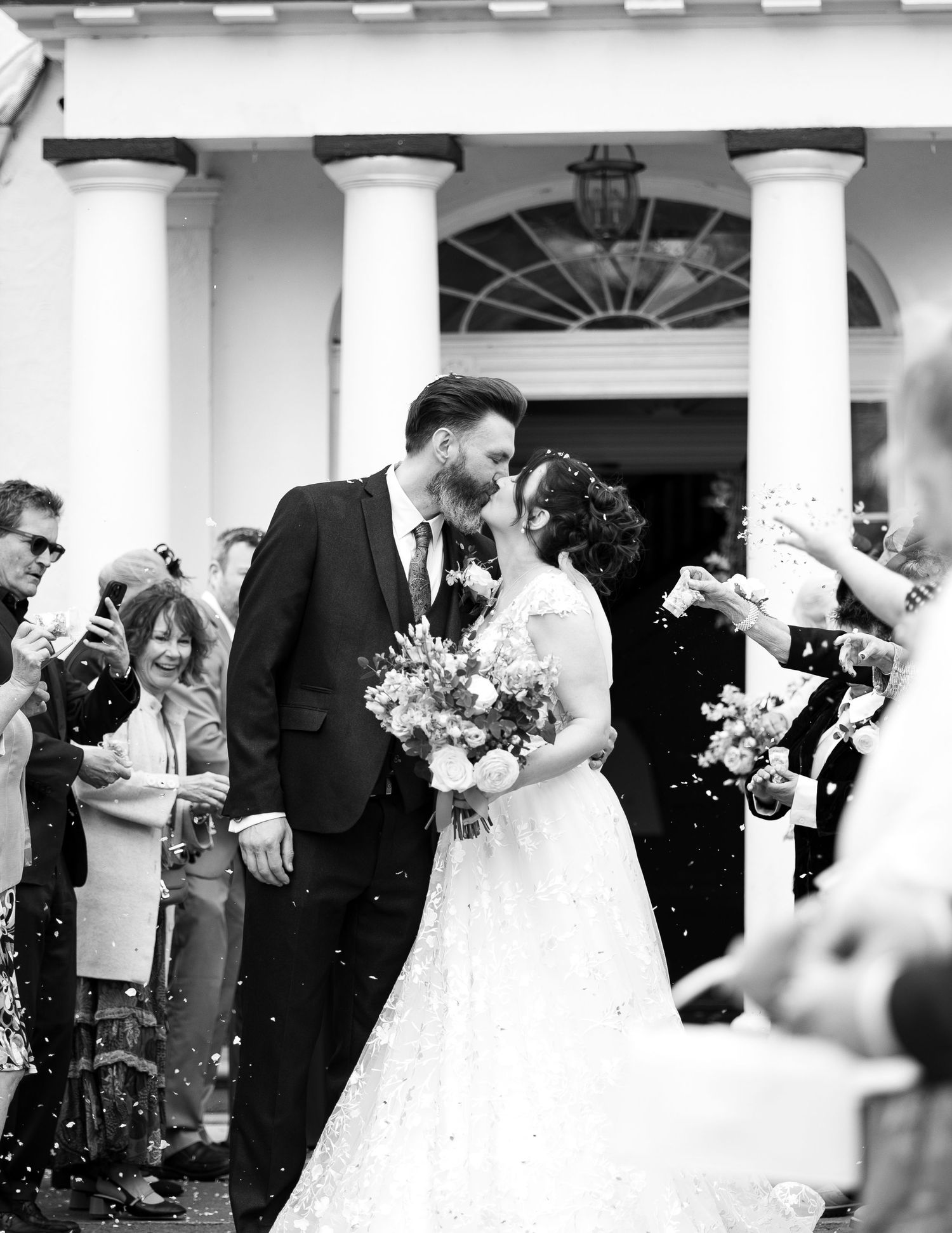 Couple kissing during confetti shot outside wedding venue. Black and white photo.