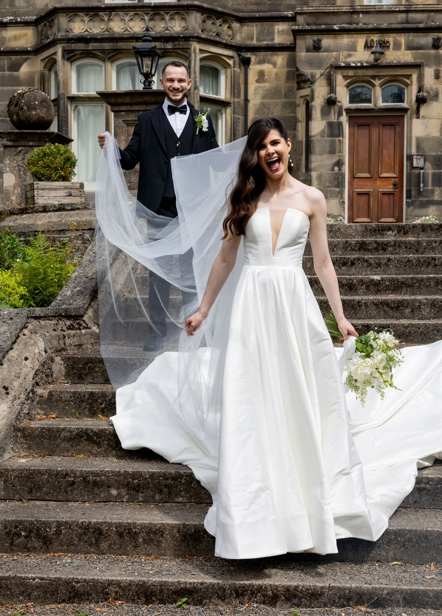 Joyous couple on the steps outside Hargate Hall. Groom holding the brides long veil. Big smiles. Natural documentary wedding photography