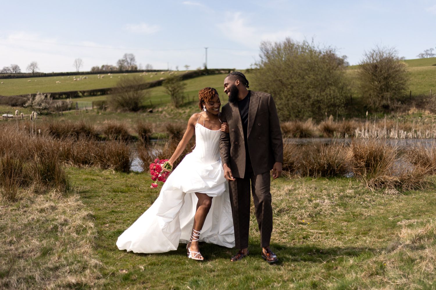Couple captured candidly hand in hand by a serene lake, carrying a bridal bouquet. Natural documentary wedding photography