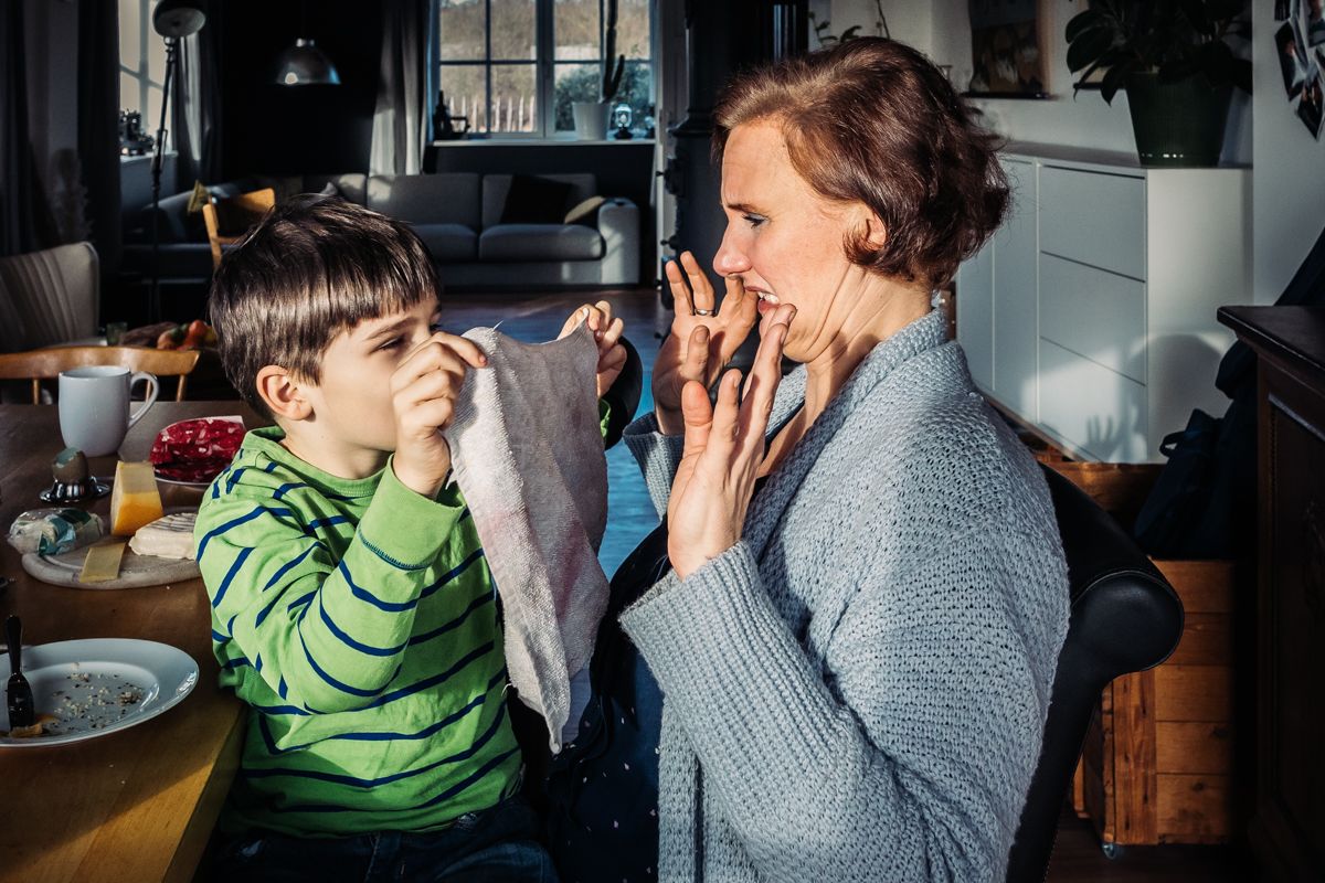 Junge hält seiner Mutter einen Spüllappen unter die Nase, beide lachen – witziger Alltagsmoment bei der Familienfotografie Lübeck