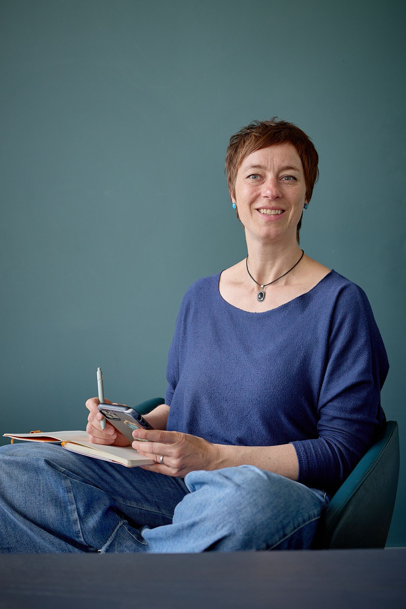 Sylvia Fischer sitzt mit Handy und Schreibzeug und schaut in die Kamera