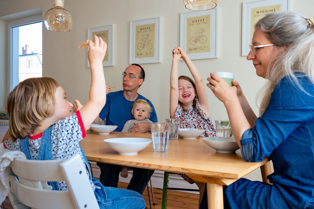 Familie isst gemeinsam am Tisch, Kinder strecken die Arme hoch – lebendiger Familienalltag bei der Familienreportage Lübeck