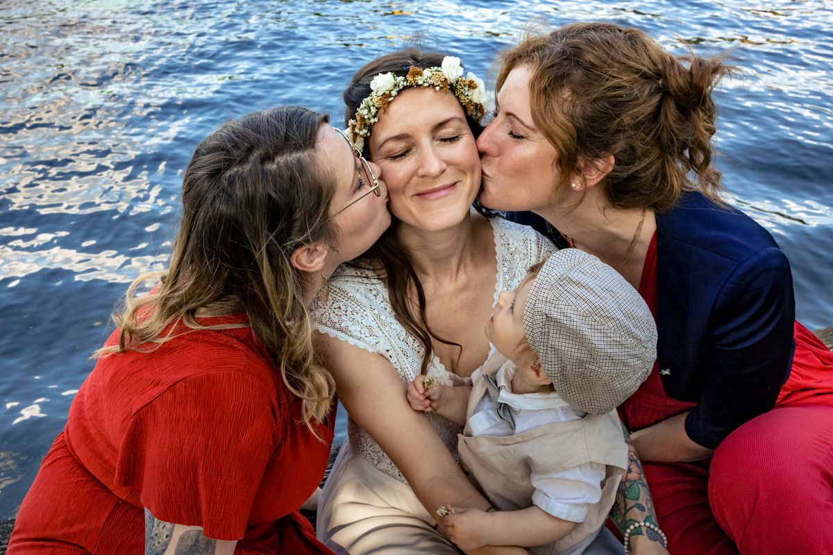 Braut wird von zwei Freundinnen geküsst, Baby auf dem Schoß – natürlicher, emotionaler Moment bei der Hochzeit – Hochzeitsfotografie Lübeck,