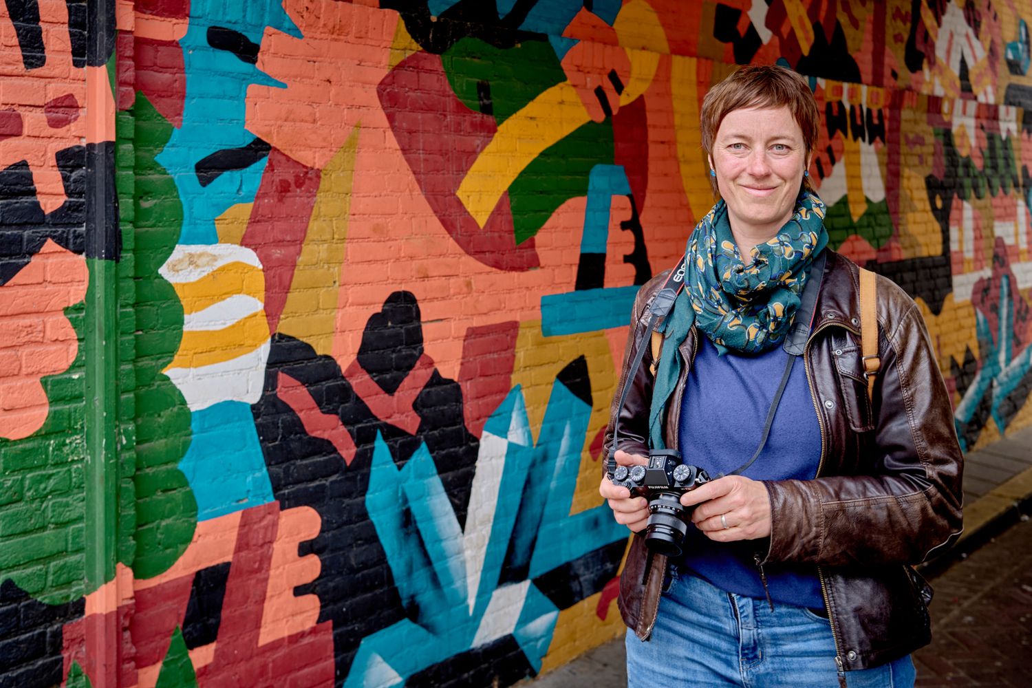 Sylvia Fischer schaut mit Kamera in der Hand vor einer bunten Grafitti-Wand in die Kamera