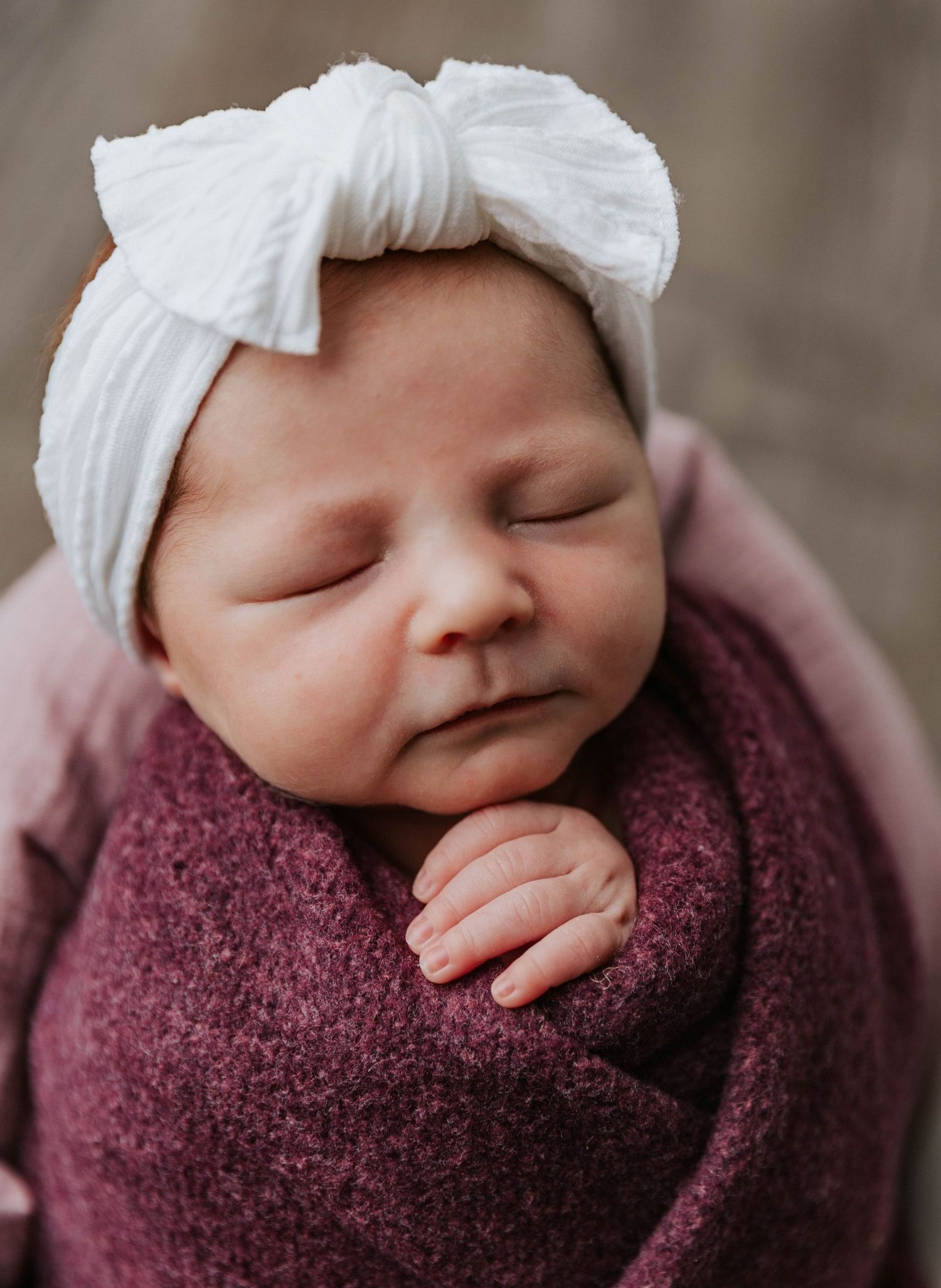 Sleeping baby wrapped in burgundy blanket wearing white bow headband in peaceful portrait.