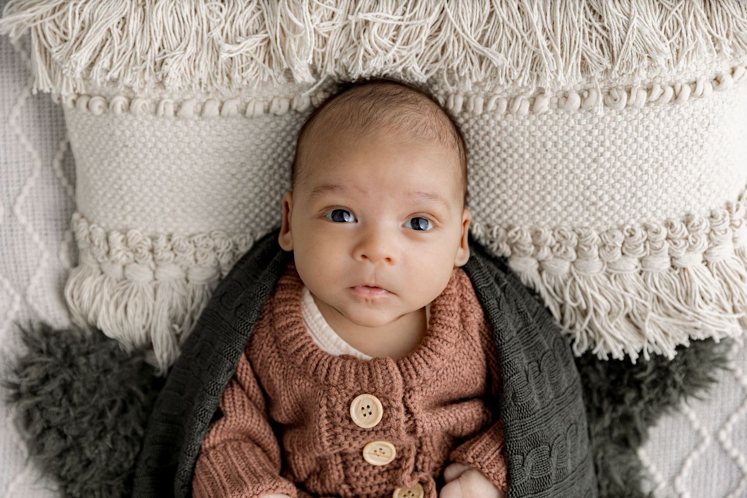 A baby wearing a brown knit sweater lies on a cream colored textured blanket with fringe details.