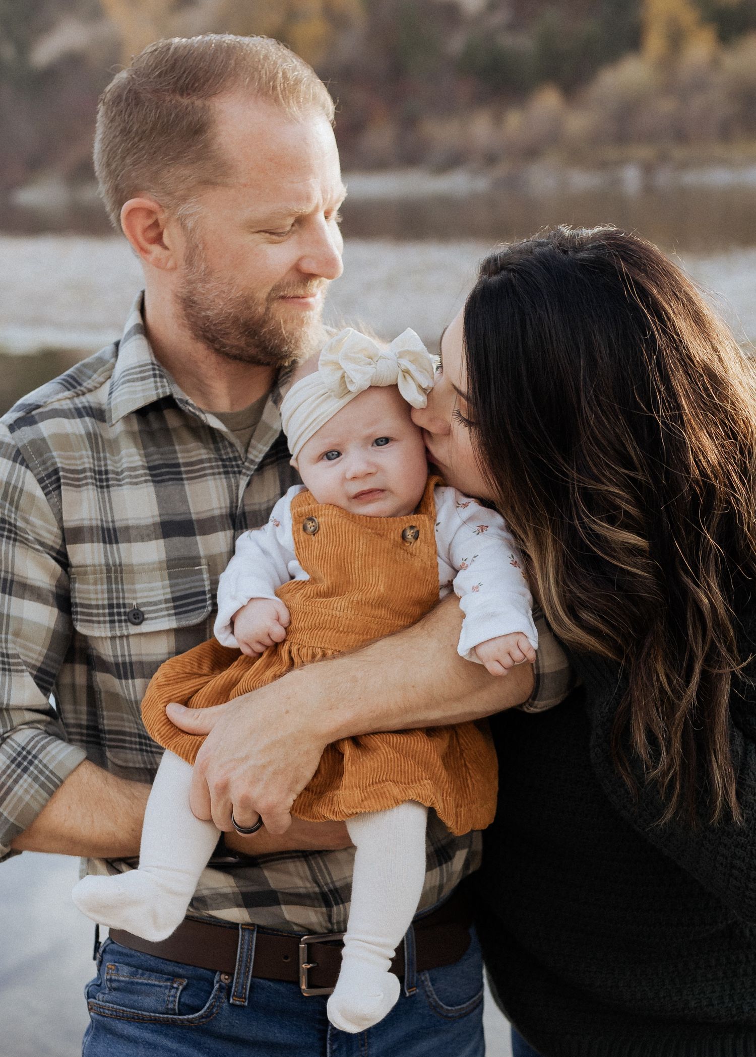 Parents sharing an intimate moment with their baby wearing brown overalls by a lake.
