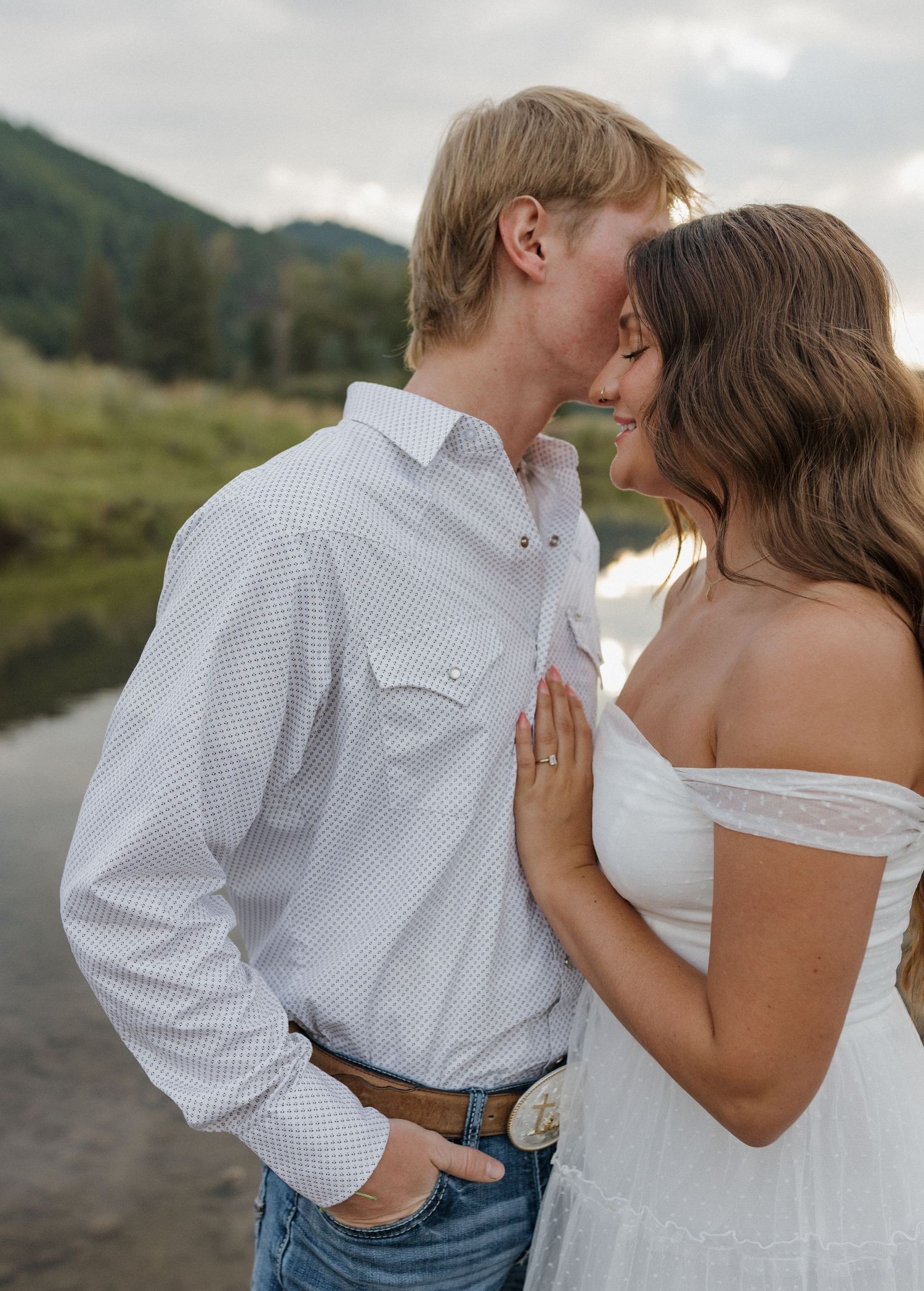 Couple sharing intimate moment by mountain lake in western attire.