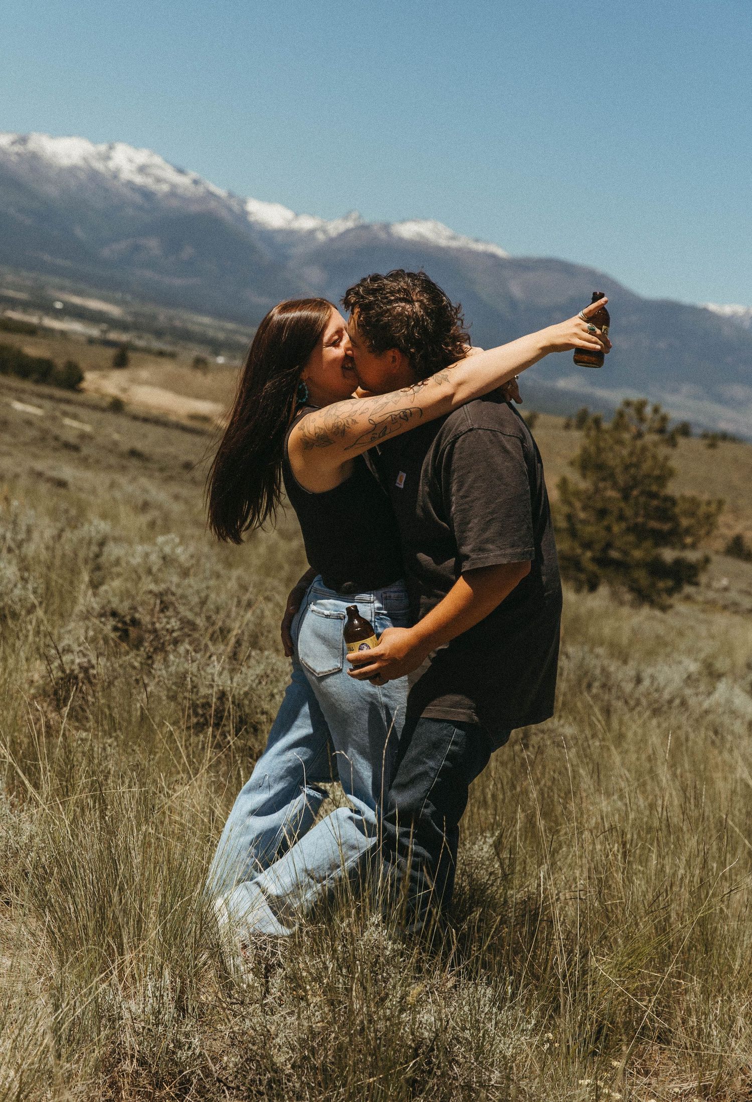 A couple shares an intimate kiss against a backdrop of snow-capped mountains and rolling hills on a sunny day.