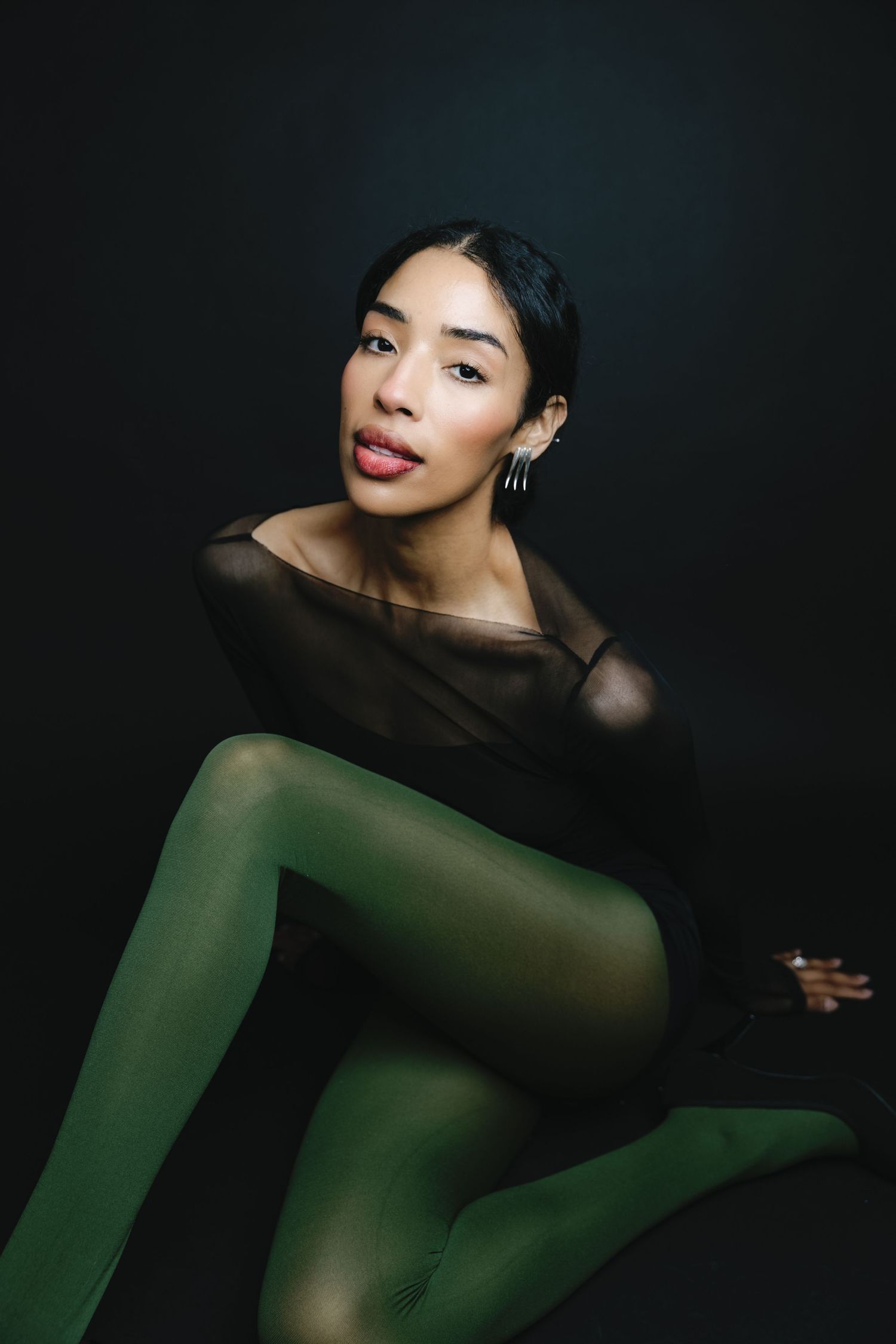 A woman in a sheer black top and green tights poses gracefully against a dark background.