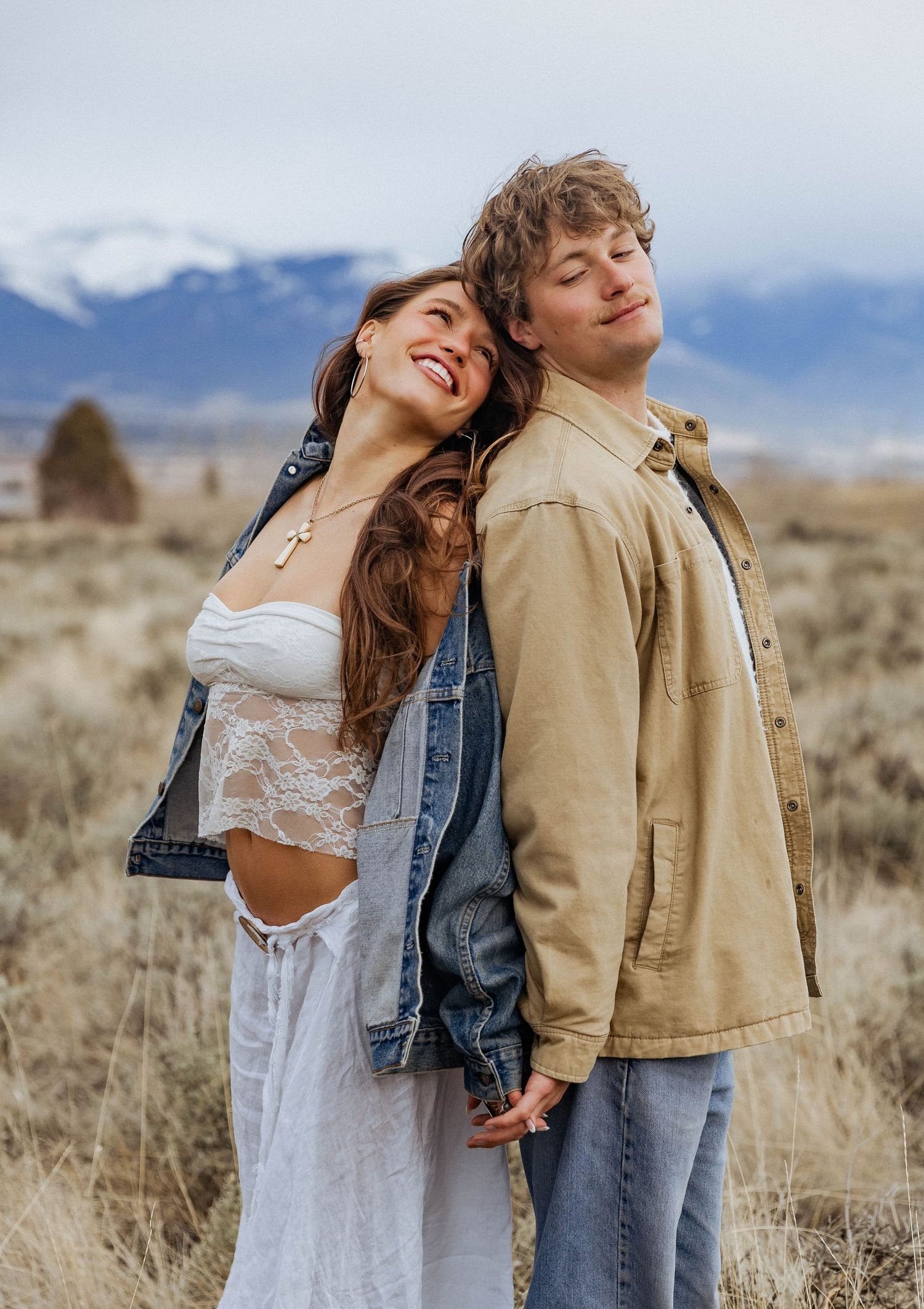 A romantic couple poses together against snow-capped mountains in casual attire.
