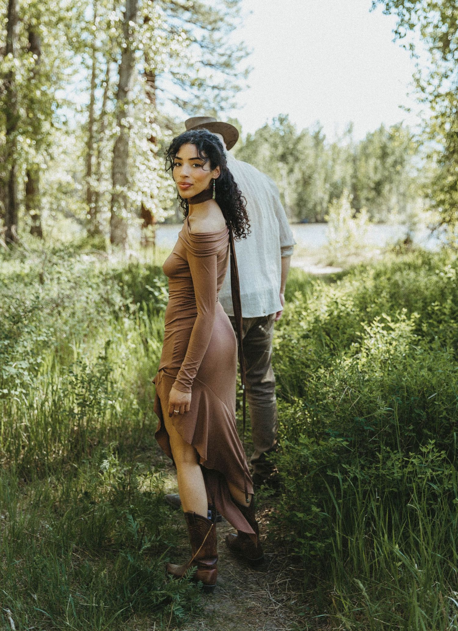 A couple embraces in a sunlit forest clearing with tall grass and pine trees creating a romantic outdoor setting.