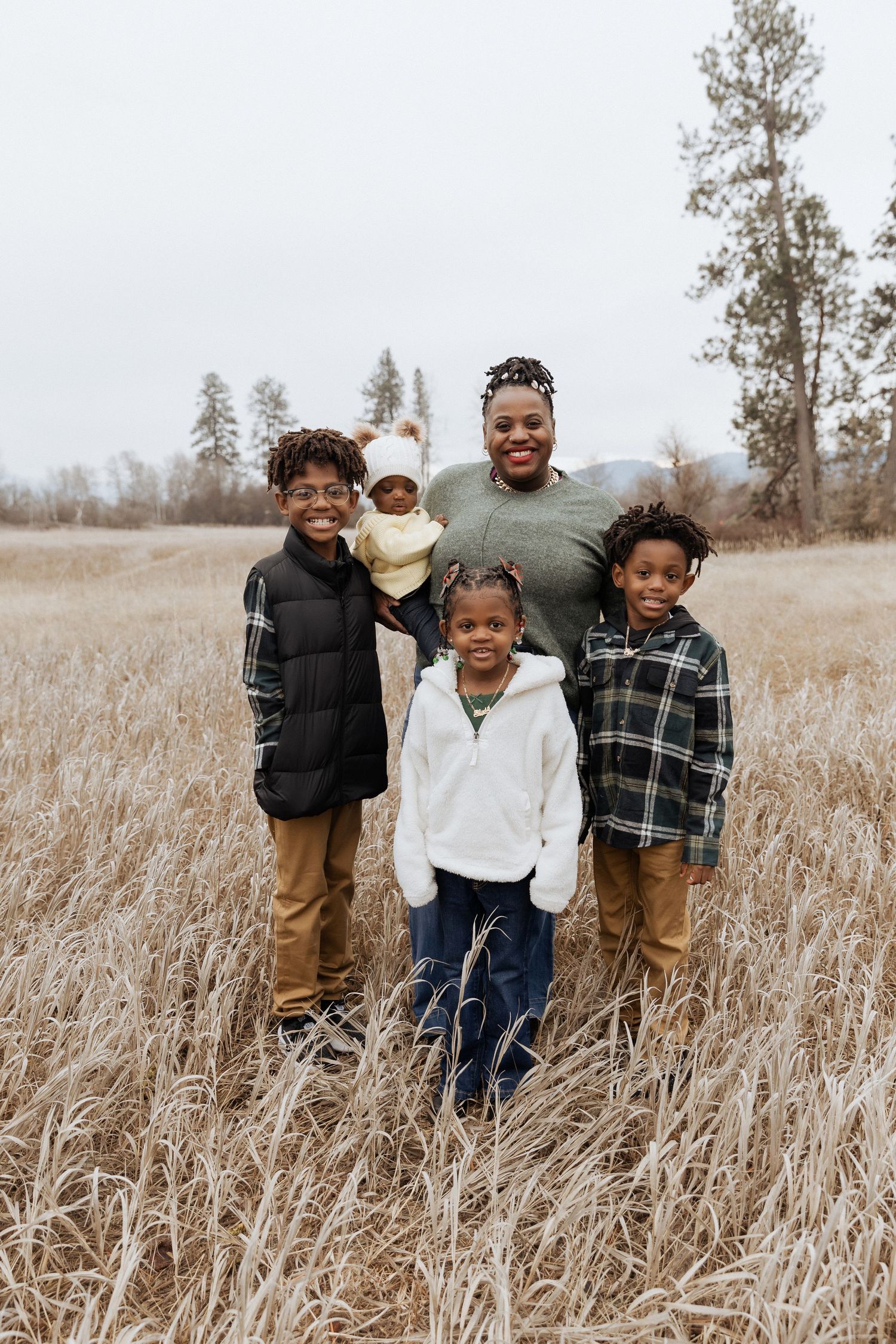 Family standing together in a golden grass field with pine trees in background.