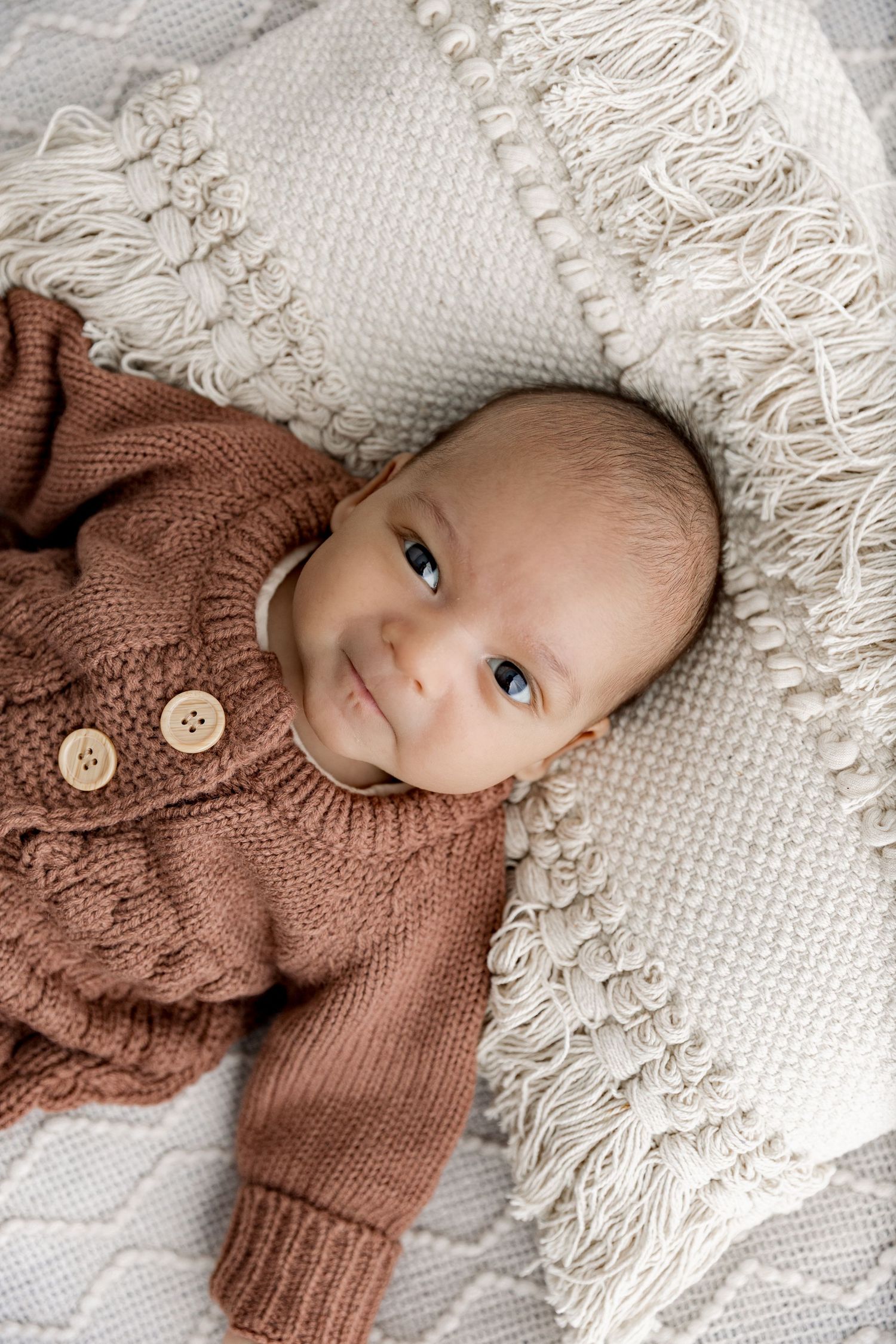 Baby in brown knit sweater lying on white textured blanket with soft neutral tones and natural lighting.