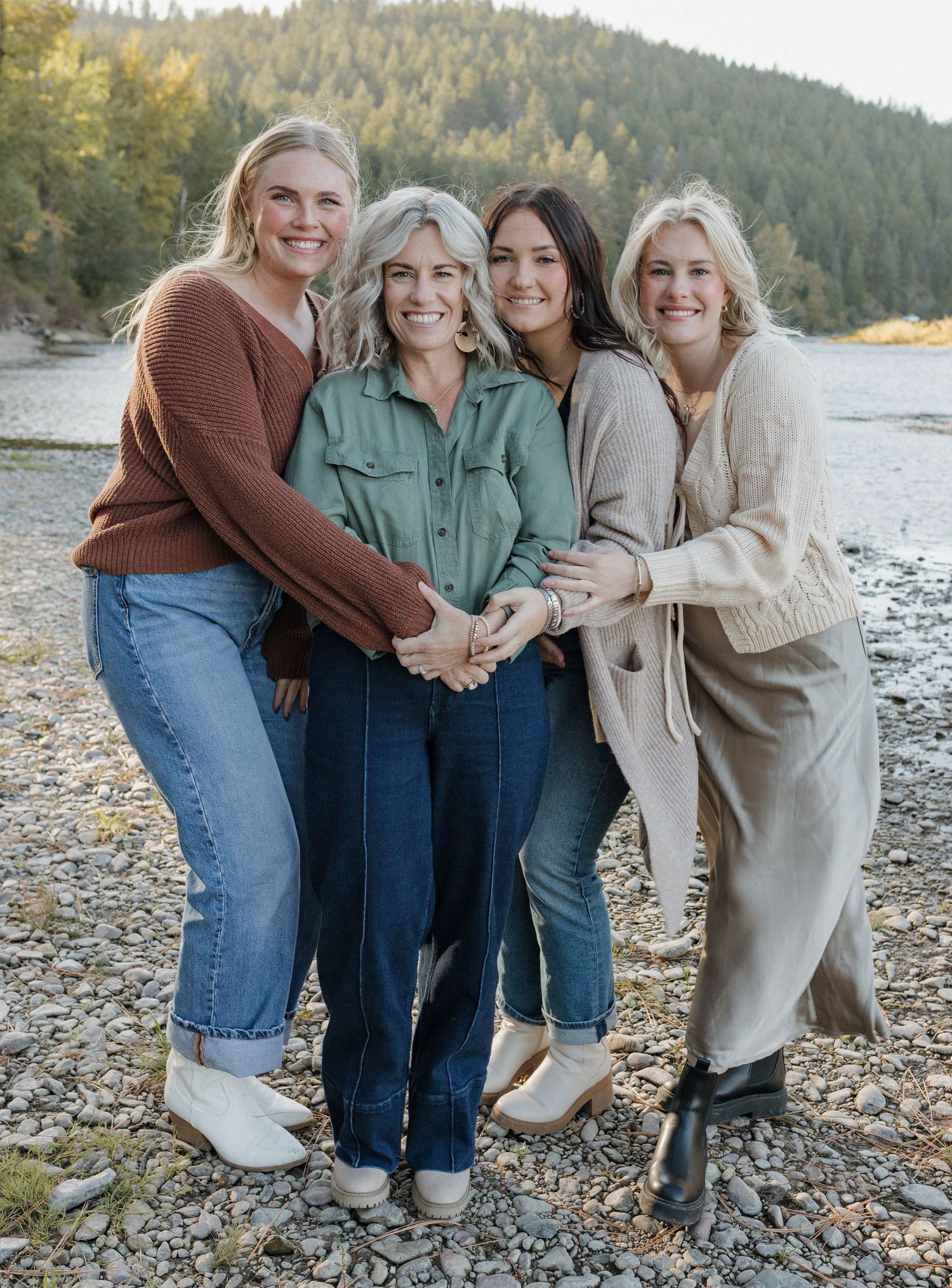 Four women embracing and smiling together on a rocky beach with evergreen forest backdrop.