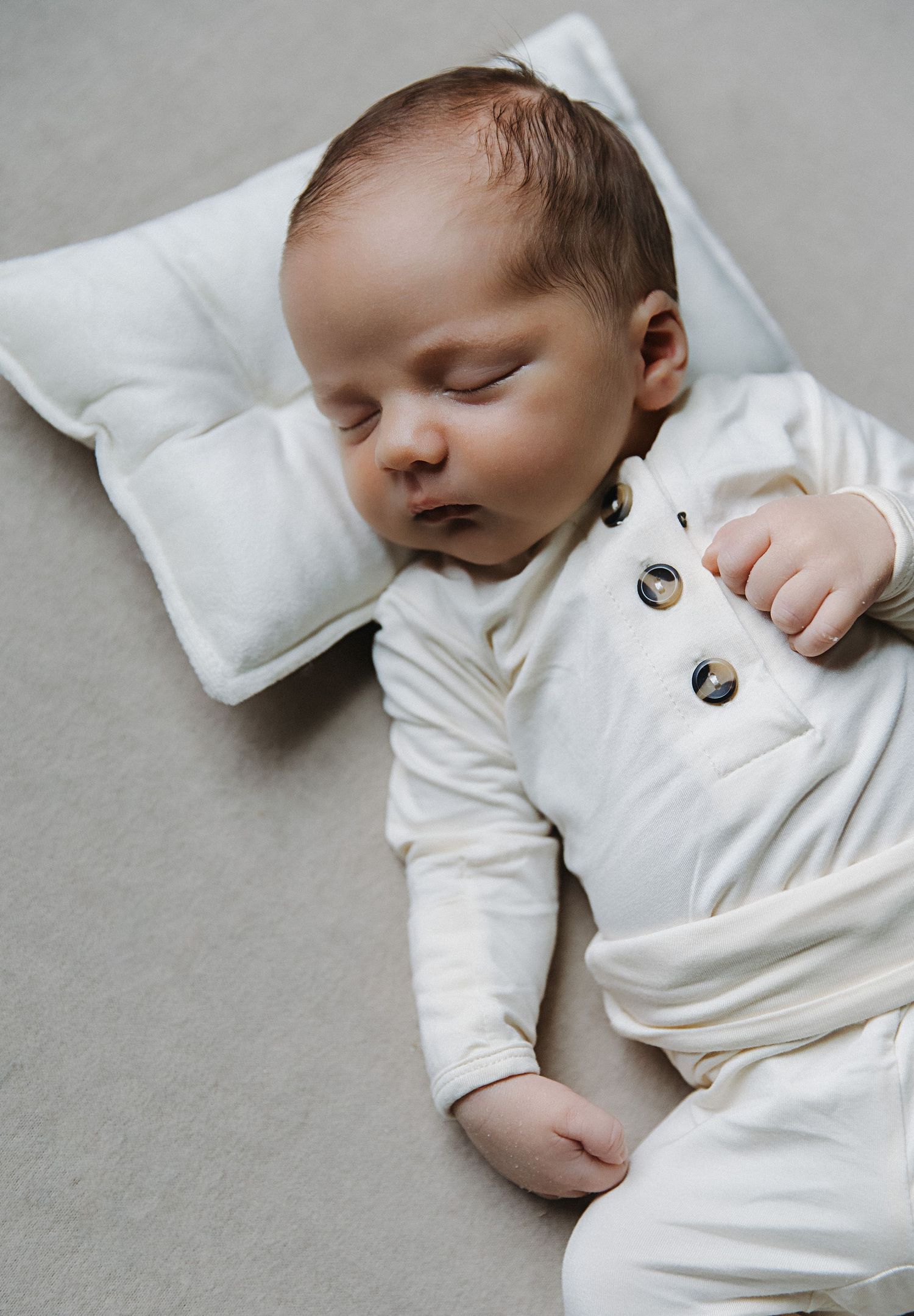 Peaceful sleeping baby wearing white button-up outfit on soft grey surface.