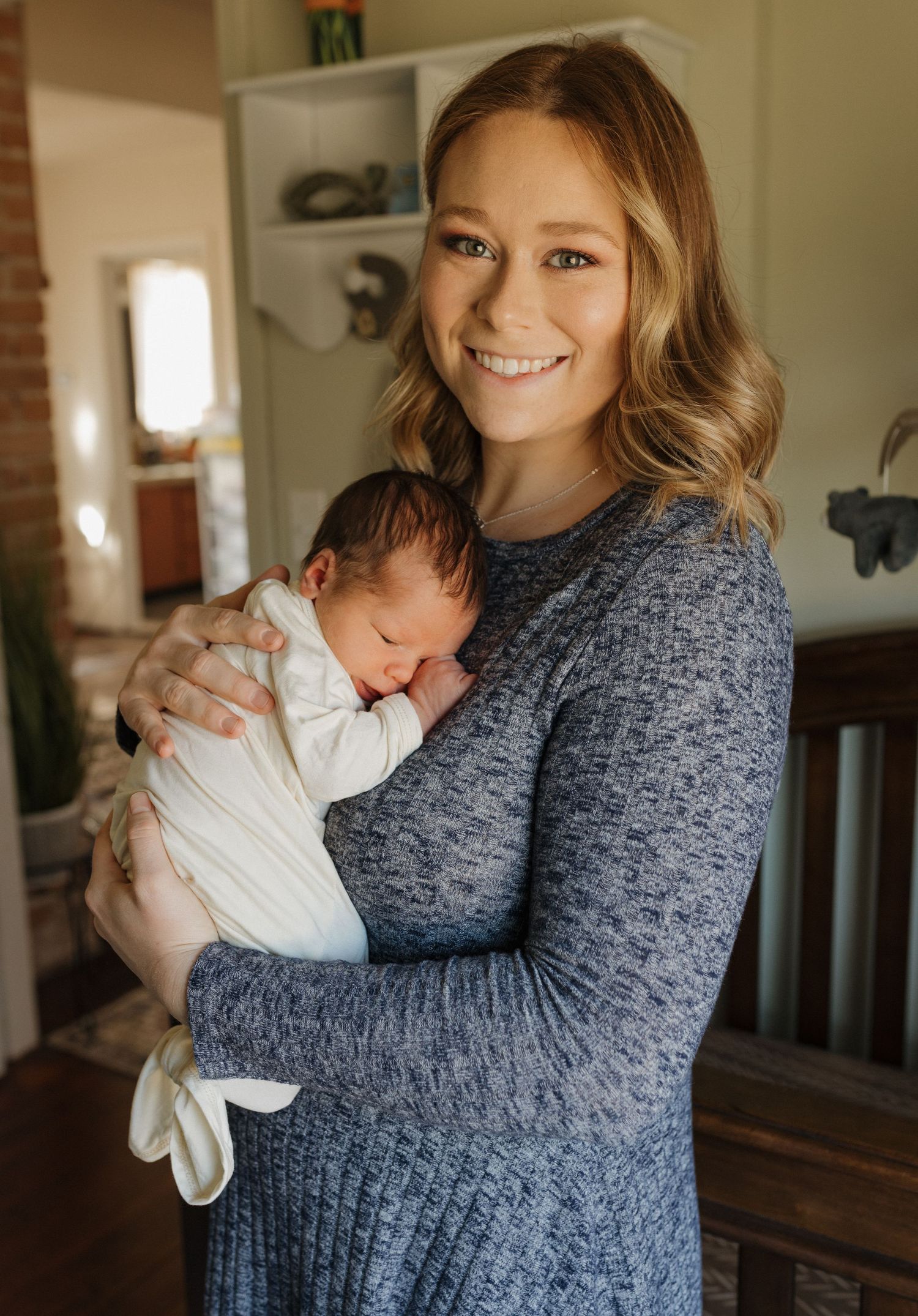 Mother in grey sweater holding sleeping newborn wrapped in white blanket.