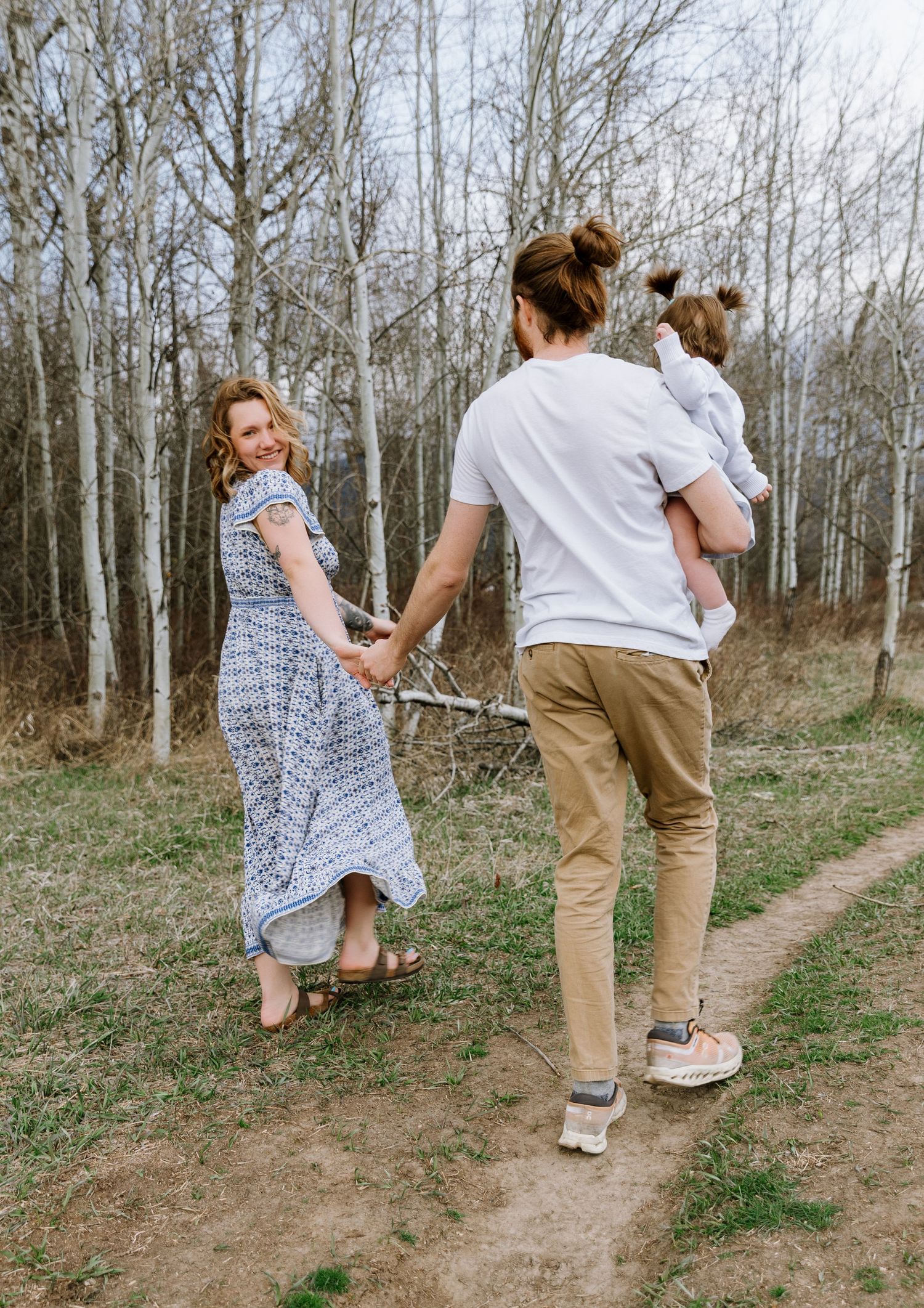 A young family walks hand-in-hand on a forest path, man carrying child, woman in blue dress looking back smiling.