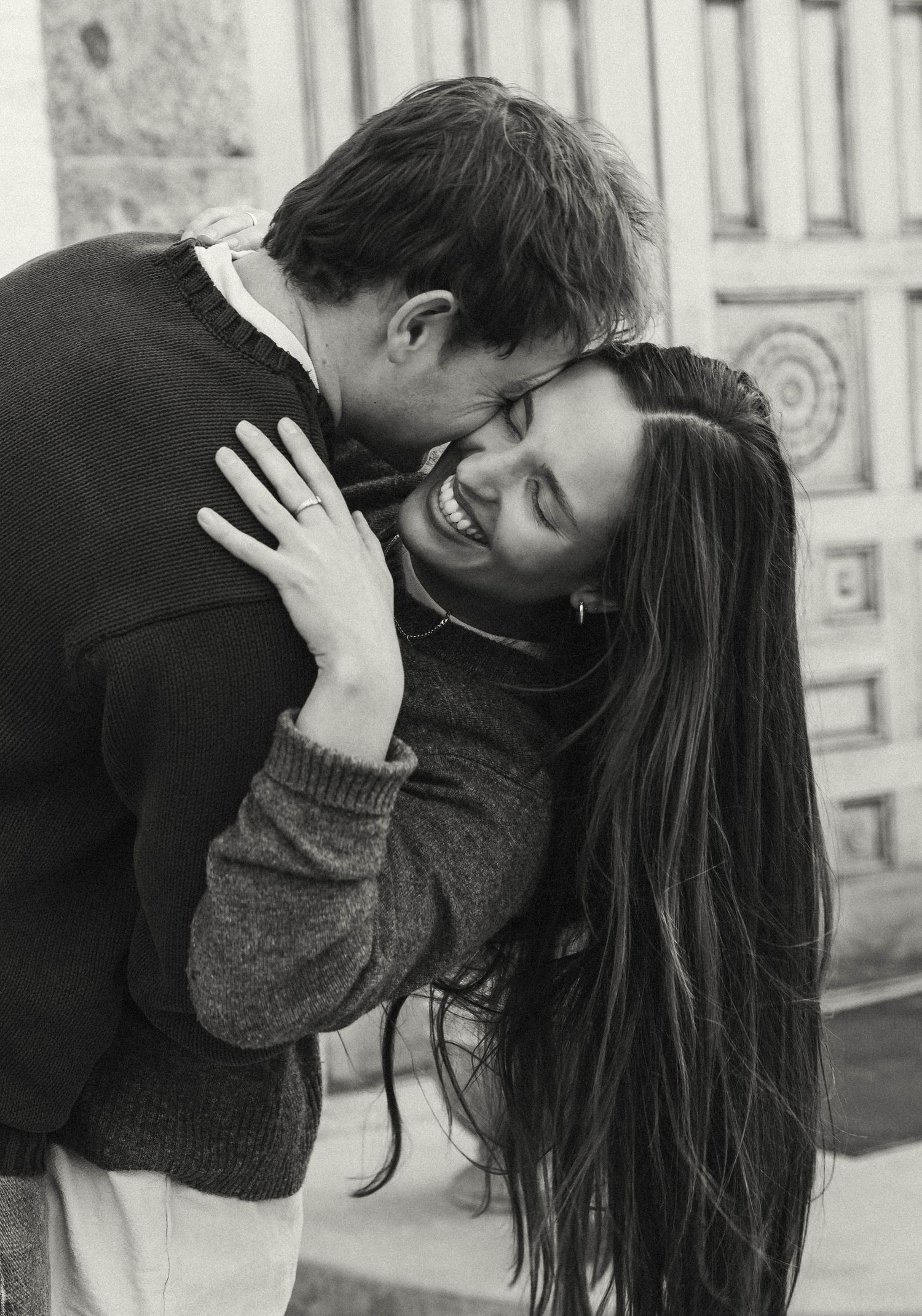A black and white portrait of a couple sharing a playful intimate moment against a stone wall.