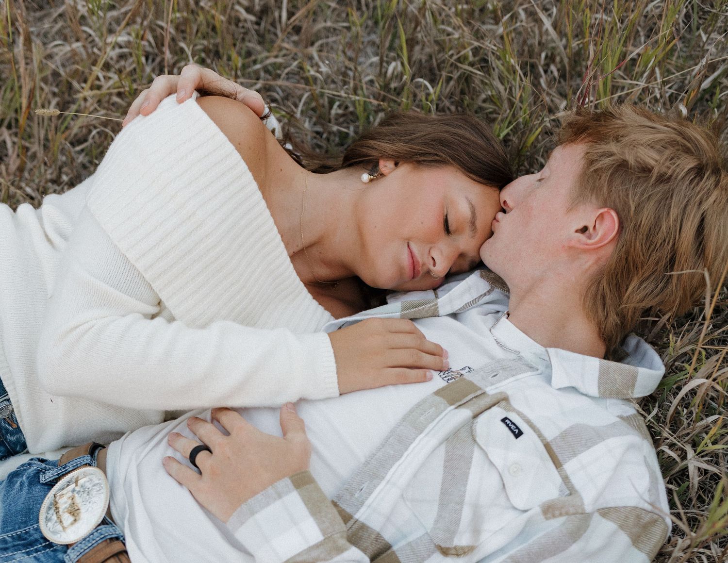 A couple in white sweaters lying together in tall grass sharing an intimate moment.
