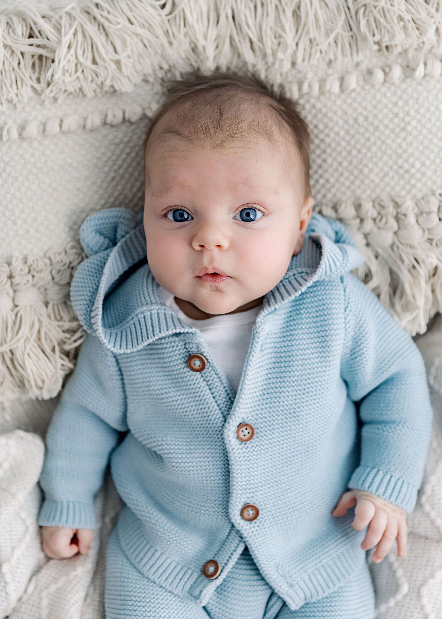 A baby wearing a light blue knit cardigan sweater with brown buttons lies on a white textured blanket.