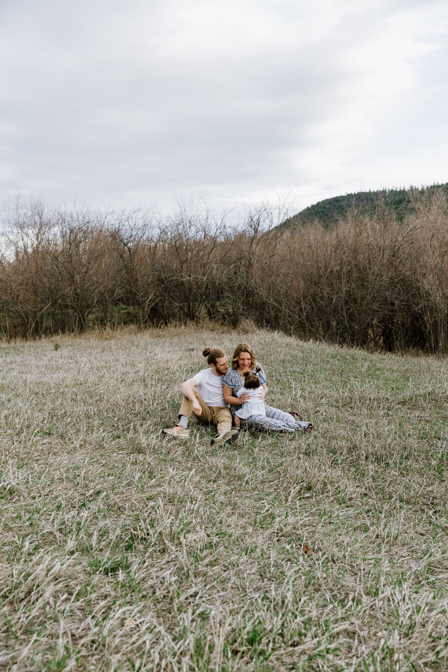 A couple sits together on a grassy field, embracing warmly with trees and a hill in the background.