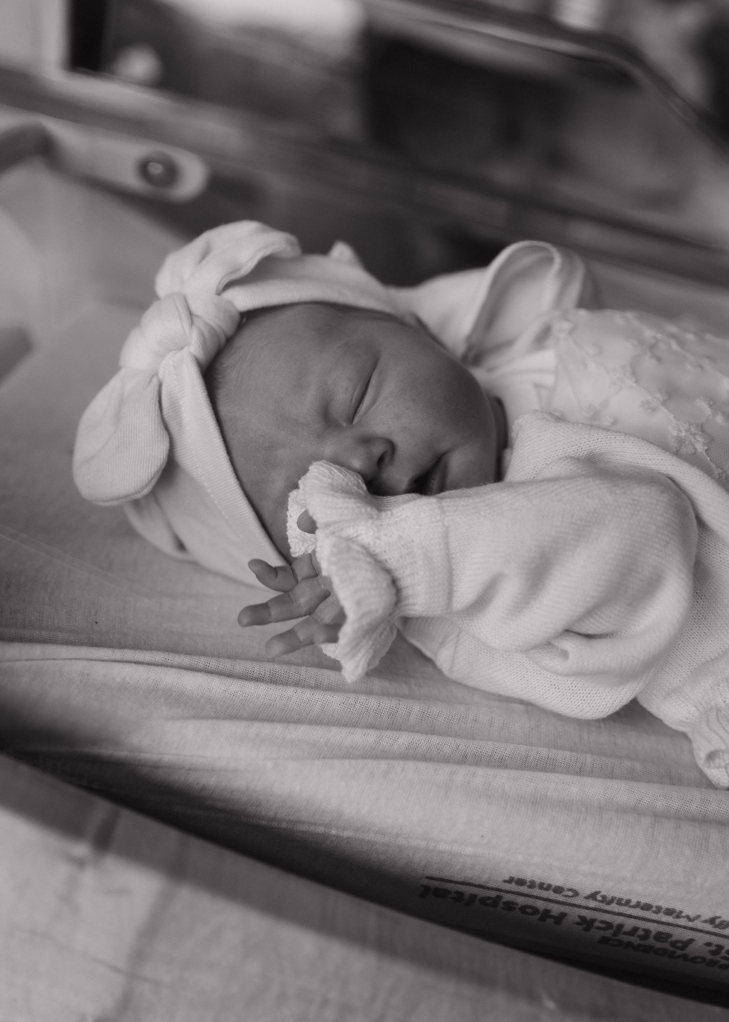 Black and white image of a sleeping newborn in white clothing and bow headband.