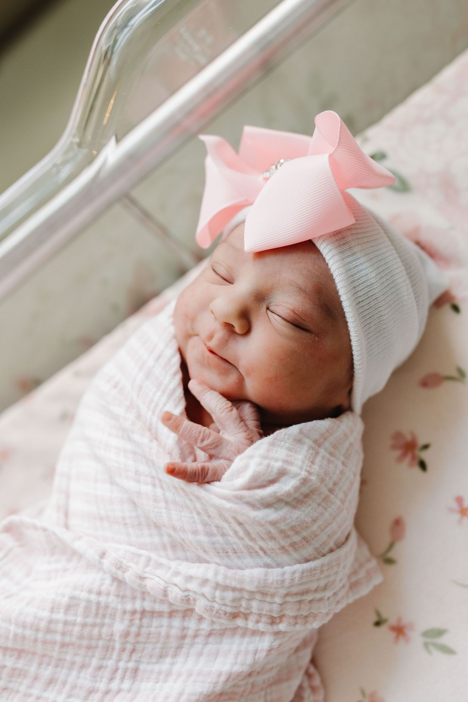 Peaceful sleeping newborn baby swaddled in white muslin wrap with pink bow headband resting on floral bedding.