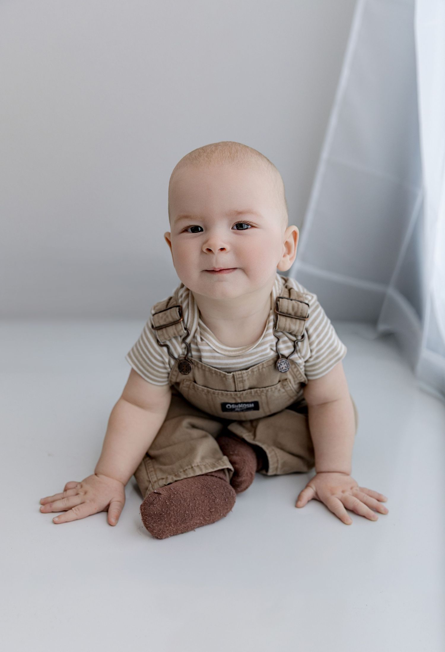 Adorable baby in striped shirt and brown overalls sitting on white floor, smiling at camera.