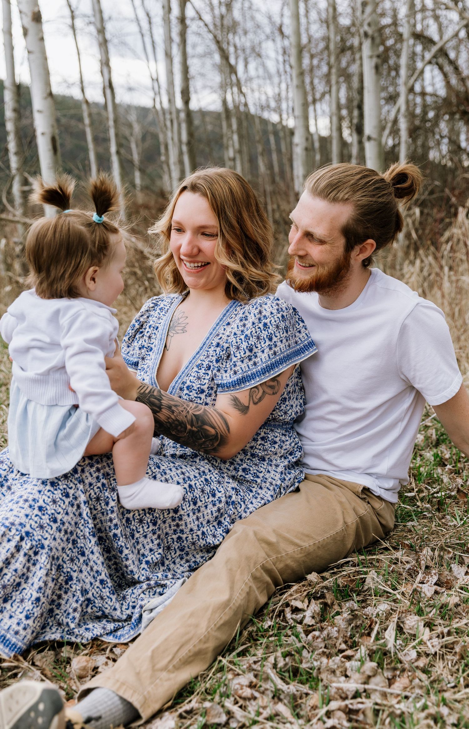 A happy young family sits outdoors among birch trees, mom in a blue floral dress holding their toddler daughter.