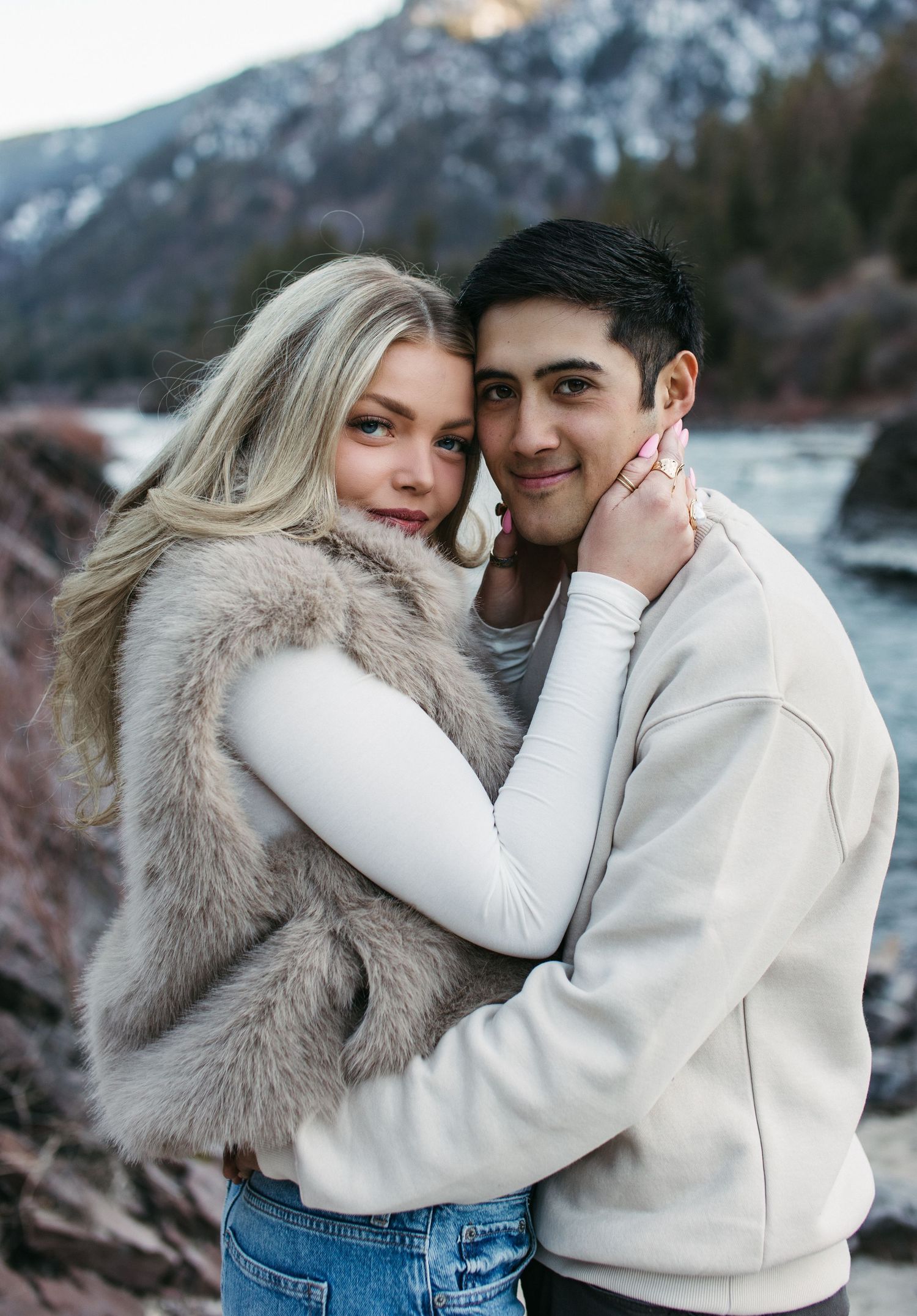 A couple in white sweaters and denim embrace outdoors near a mountainous landscape during winter.