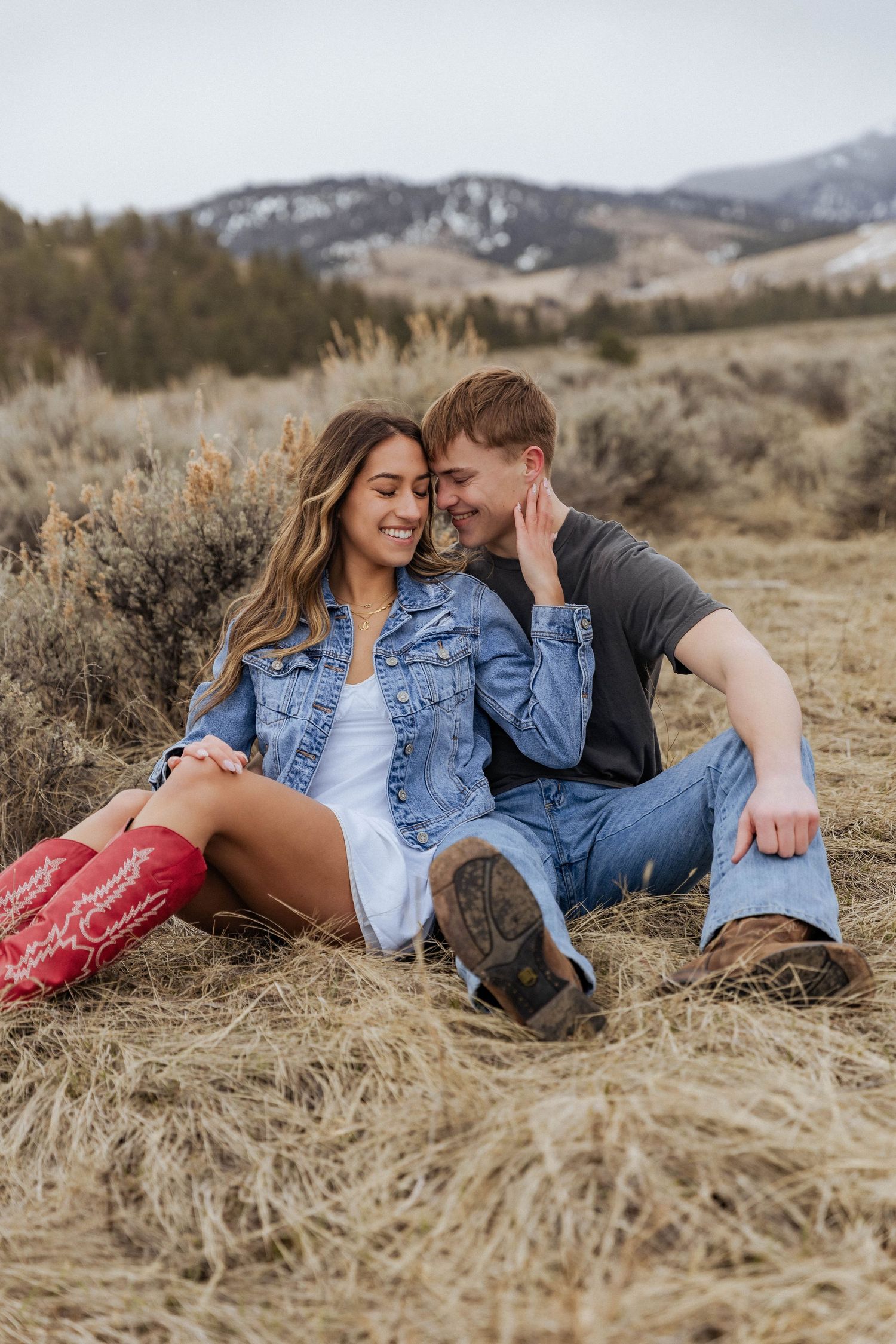 A couple sharing an intimate moment while sitting in dry grass with mountains in the background.