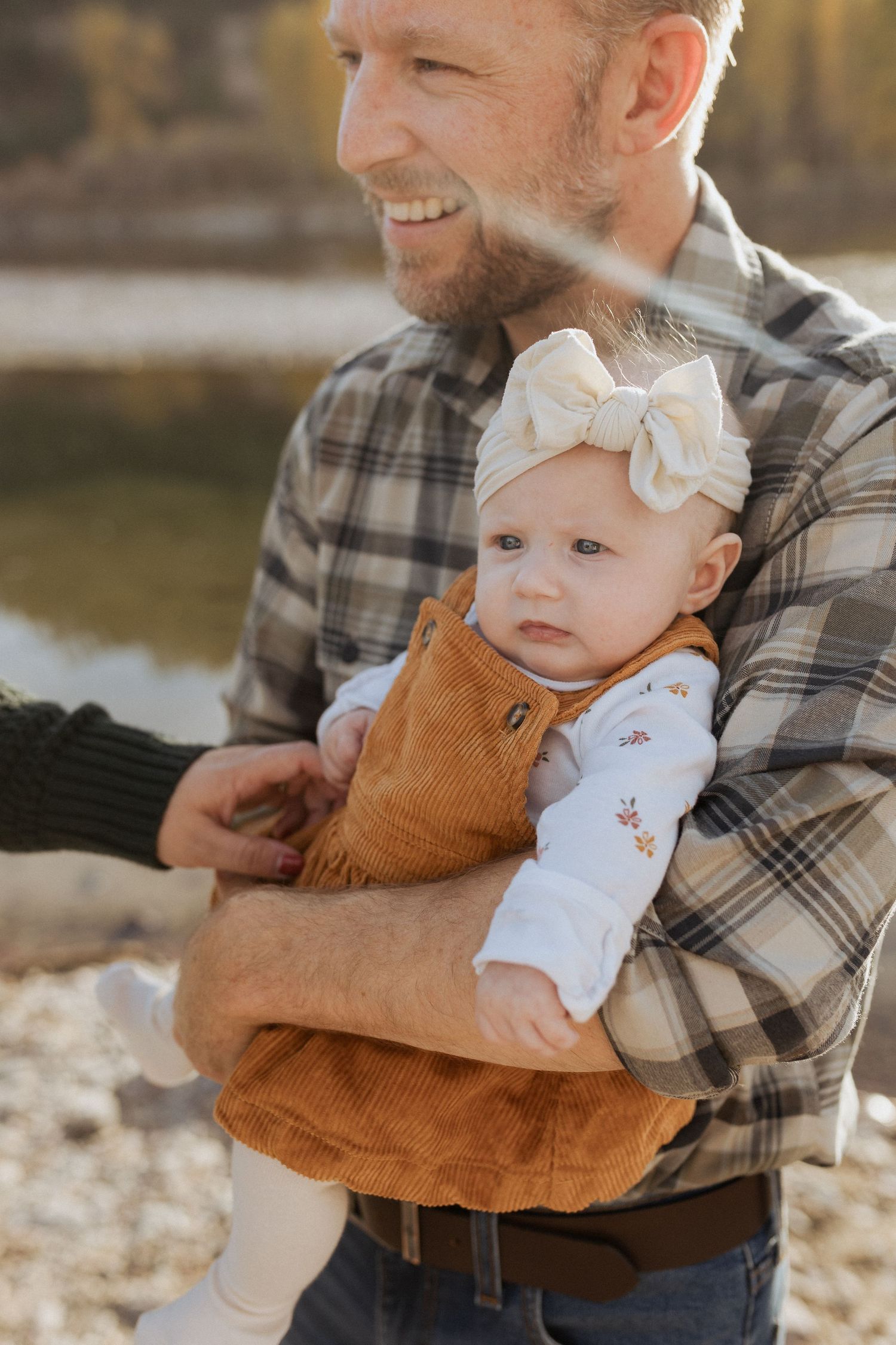Baby in corduroy overalls and bow smiling with parent by lake.