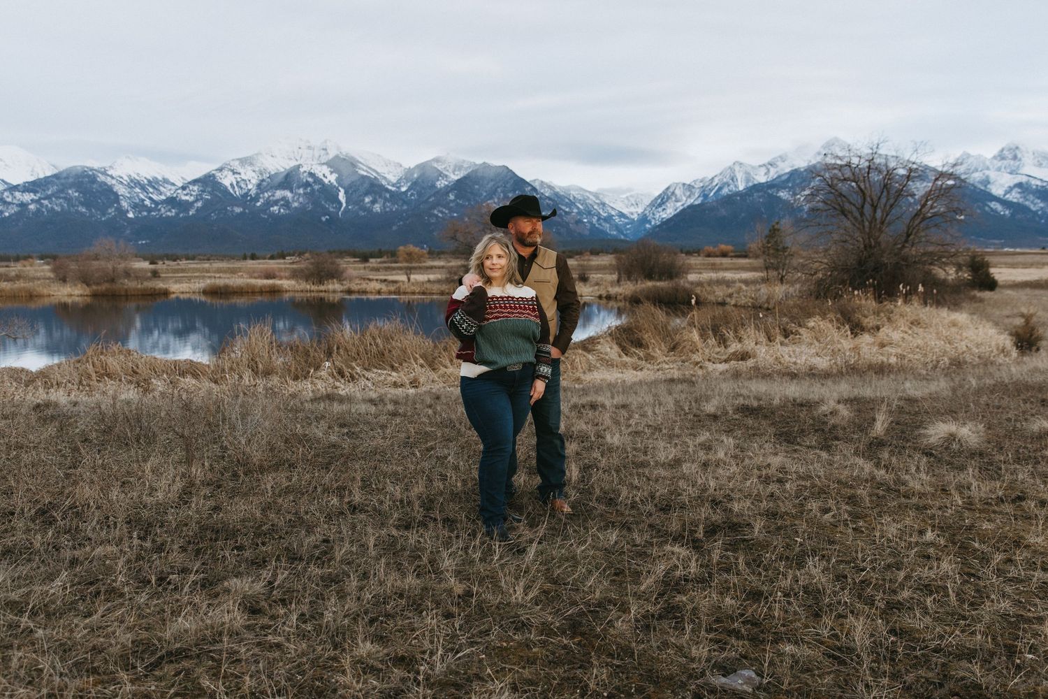 Couple embraces in a scenic mountain landscape with snow-capped peaks and dry grass field in the foreground.