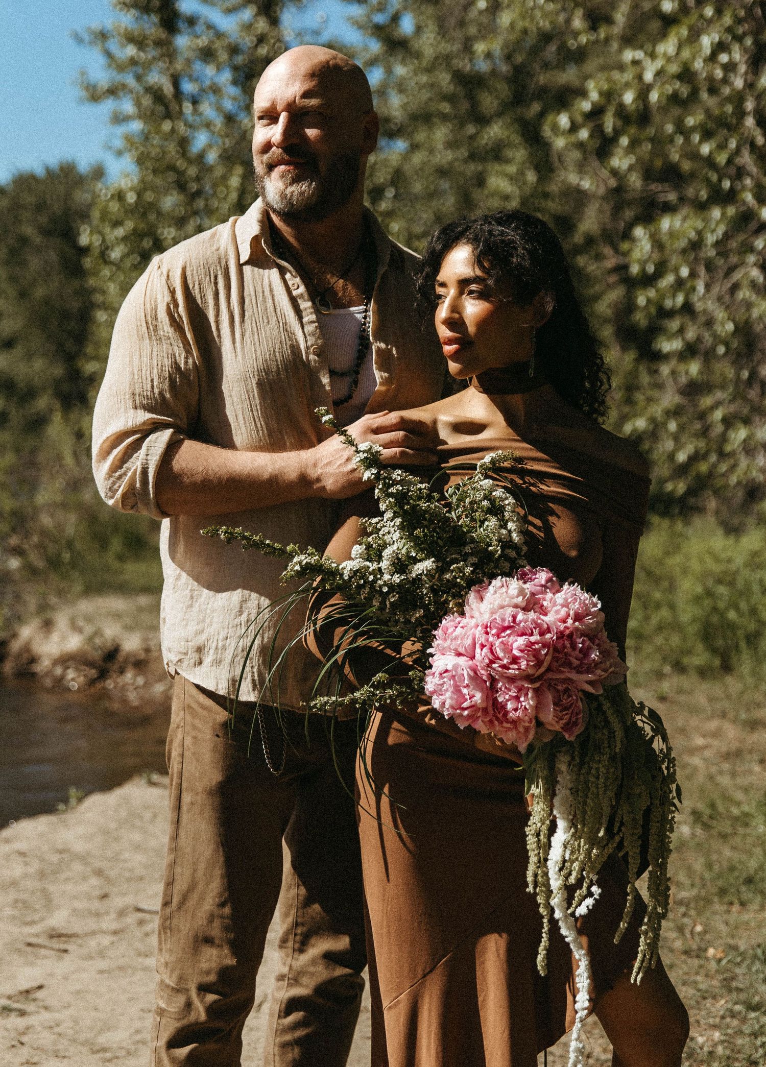 A couple embraces outdoors while holding a beautiful bouquet of pink peonies and cascading greenery on a sunny day.