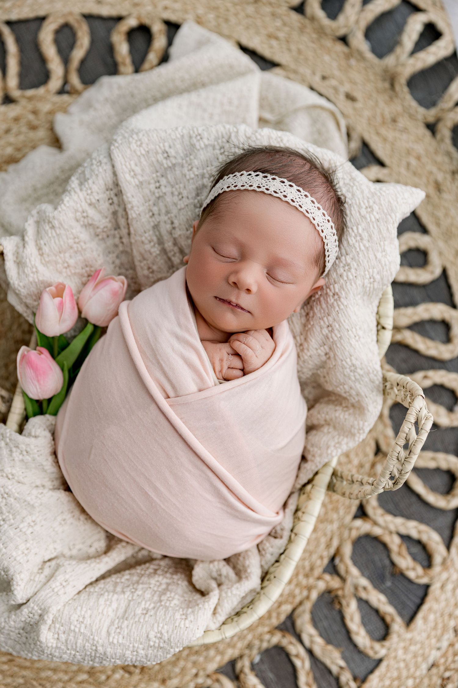 Sleeping newborn baby swaddled in pink wrap with lace headband, resting in wicker basket with tulips.