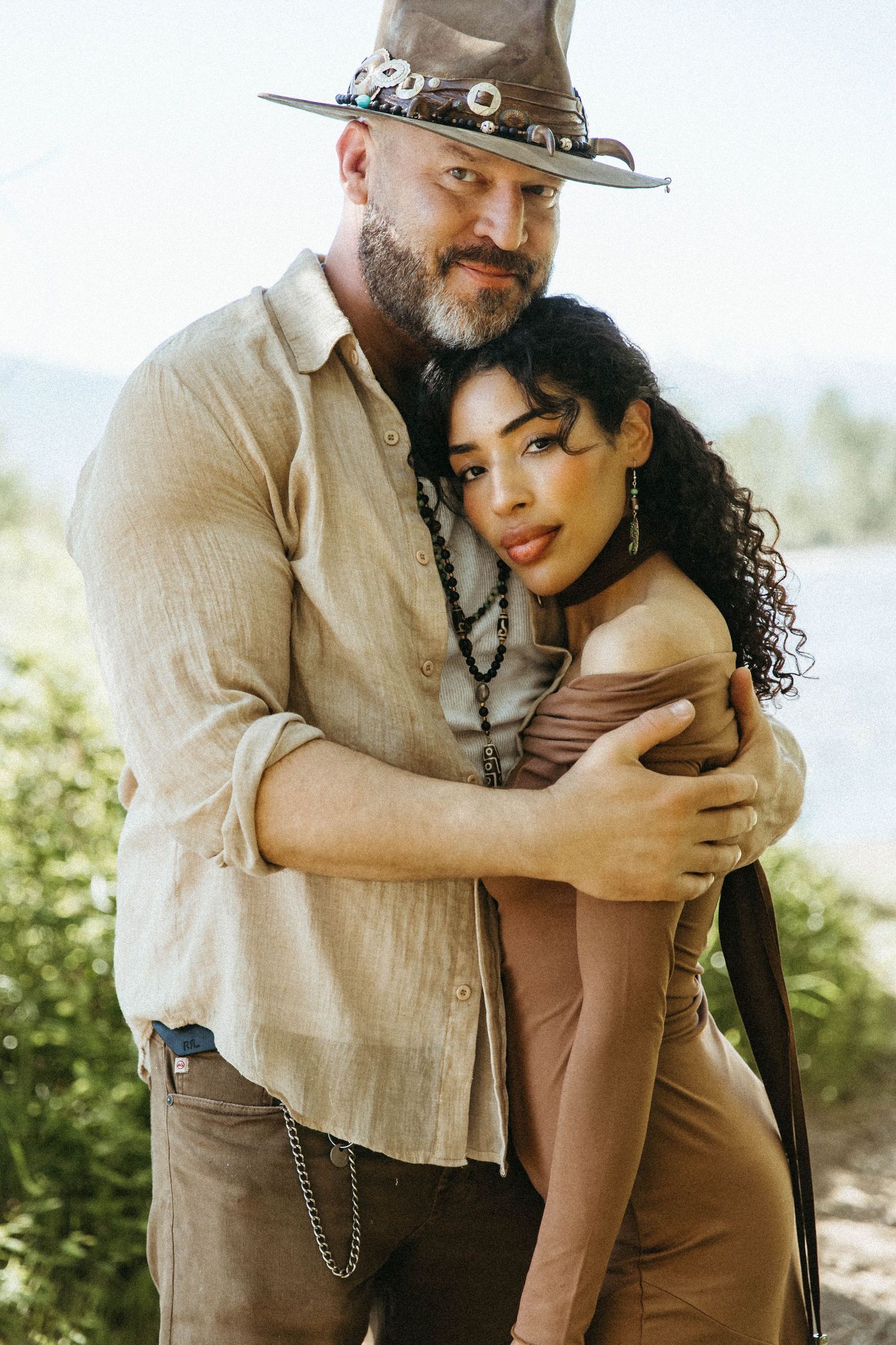 A romantic couple embrace in an outdoor setting wearing neutral toned clothing in sunlit natural surroundings.