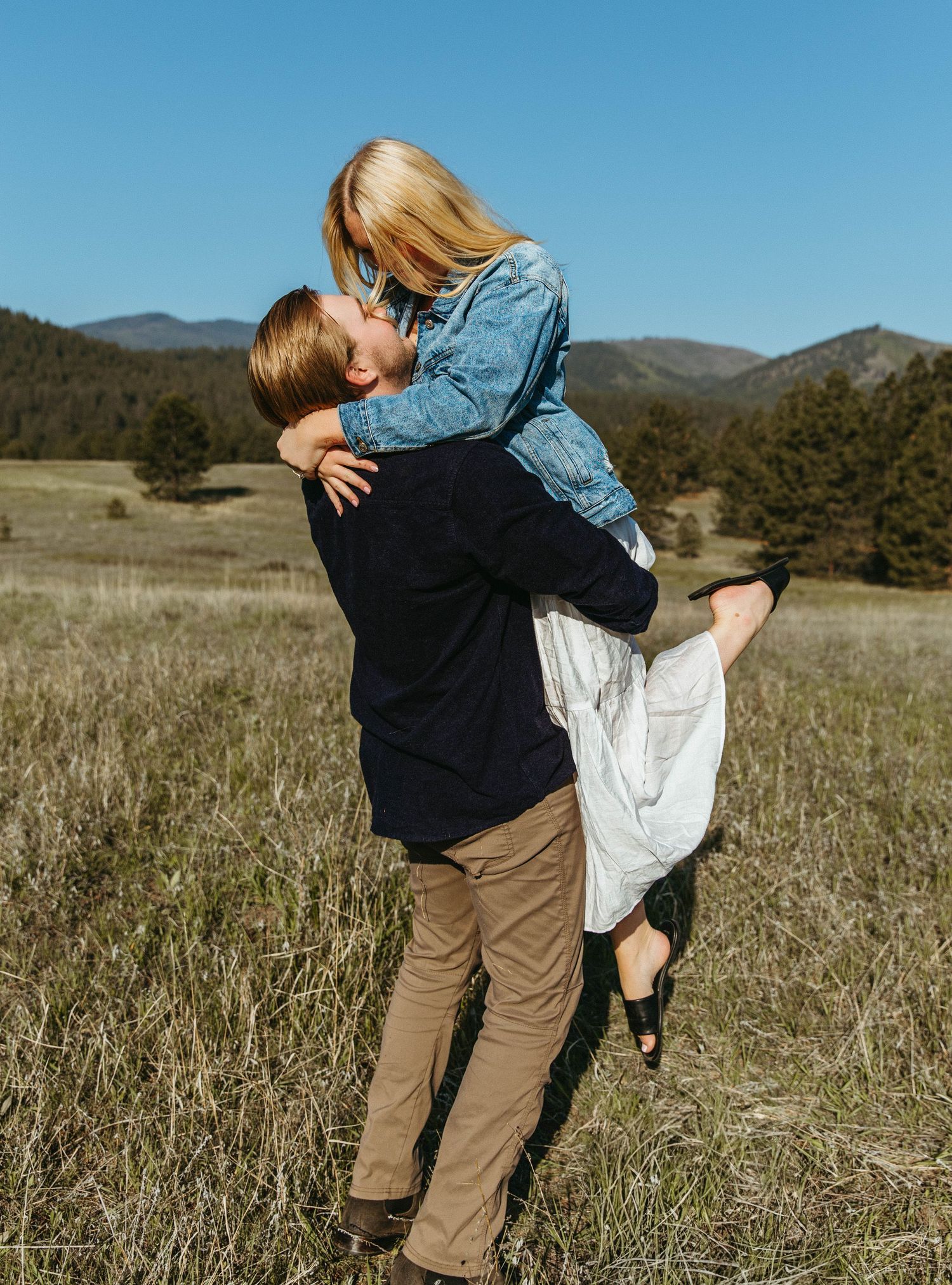 A couple embraces playfully in a sun-drenched meadow with mountains visible in the background.